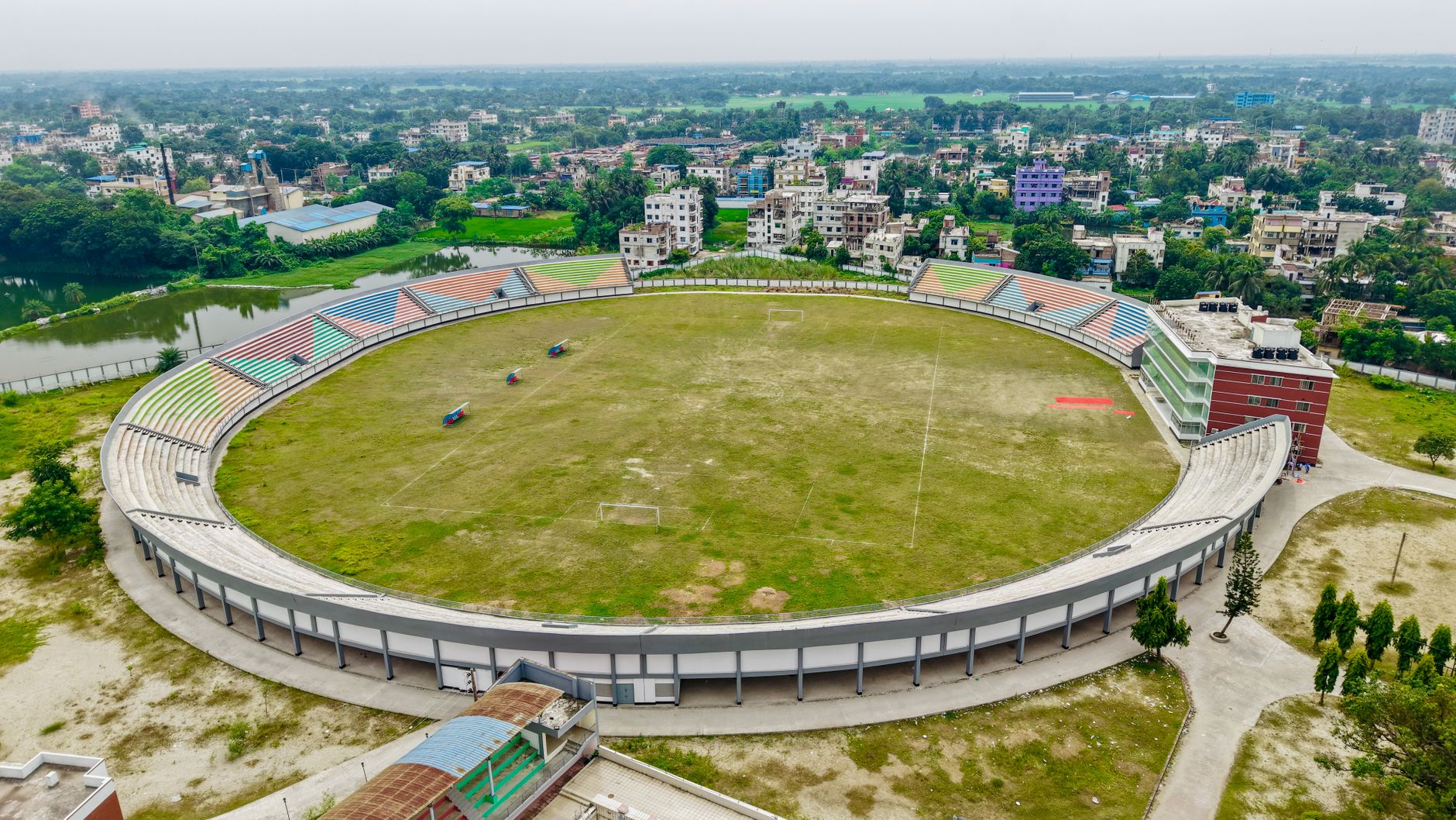 a wide aerial view of a large modern stadium exterior surrounded by security perimeter fencing and logistics vehicles during daytime, no people as focal point
