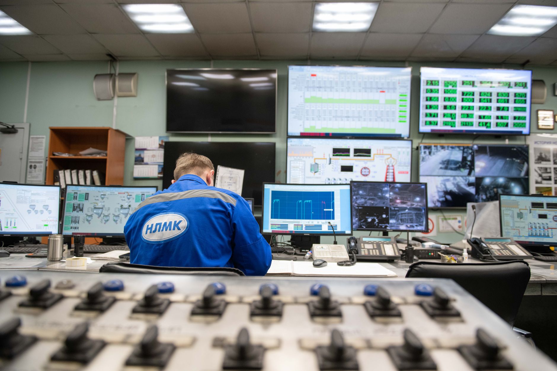 a ground control station interior at an air base, rows of flat-panel displays showing aircraft telemetry data, no personnel as focal point, equipment and screens as the primary subject in dim blue lighting
