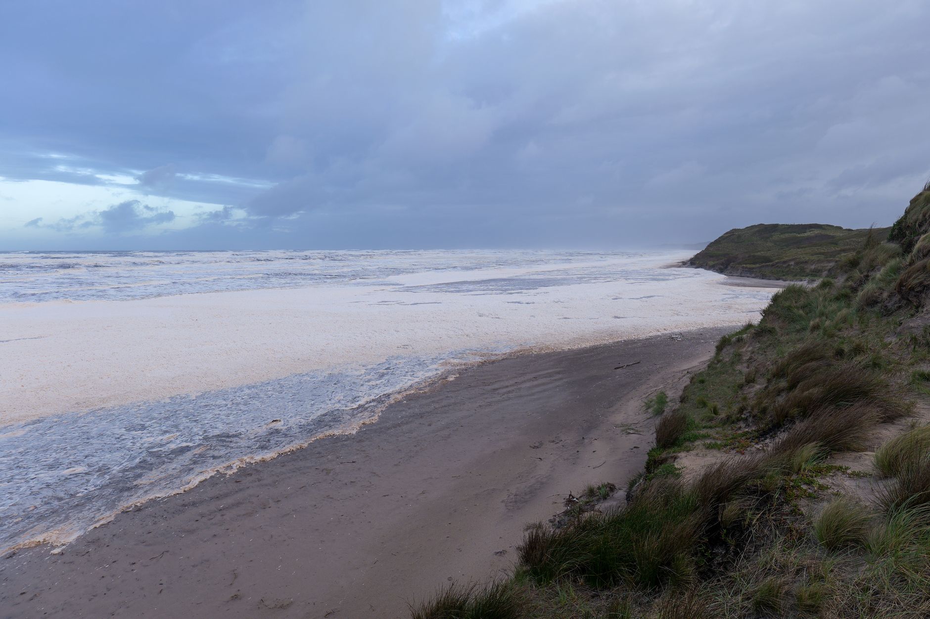 wide aerial view of a curved sandy beach in Normandy at low tide, with concrete bunker remnants visible on a grassy bluff above the shoreline