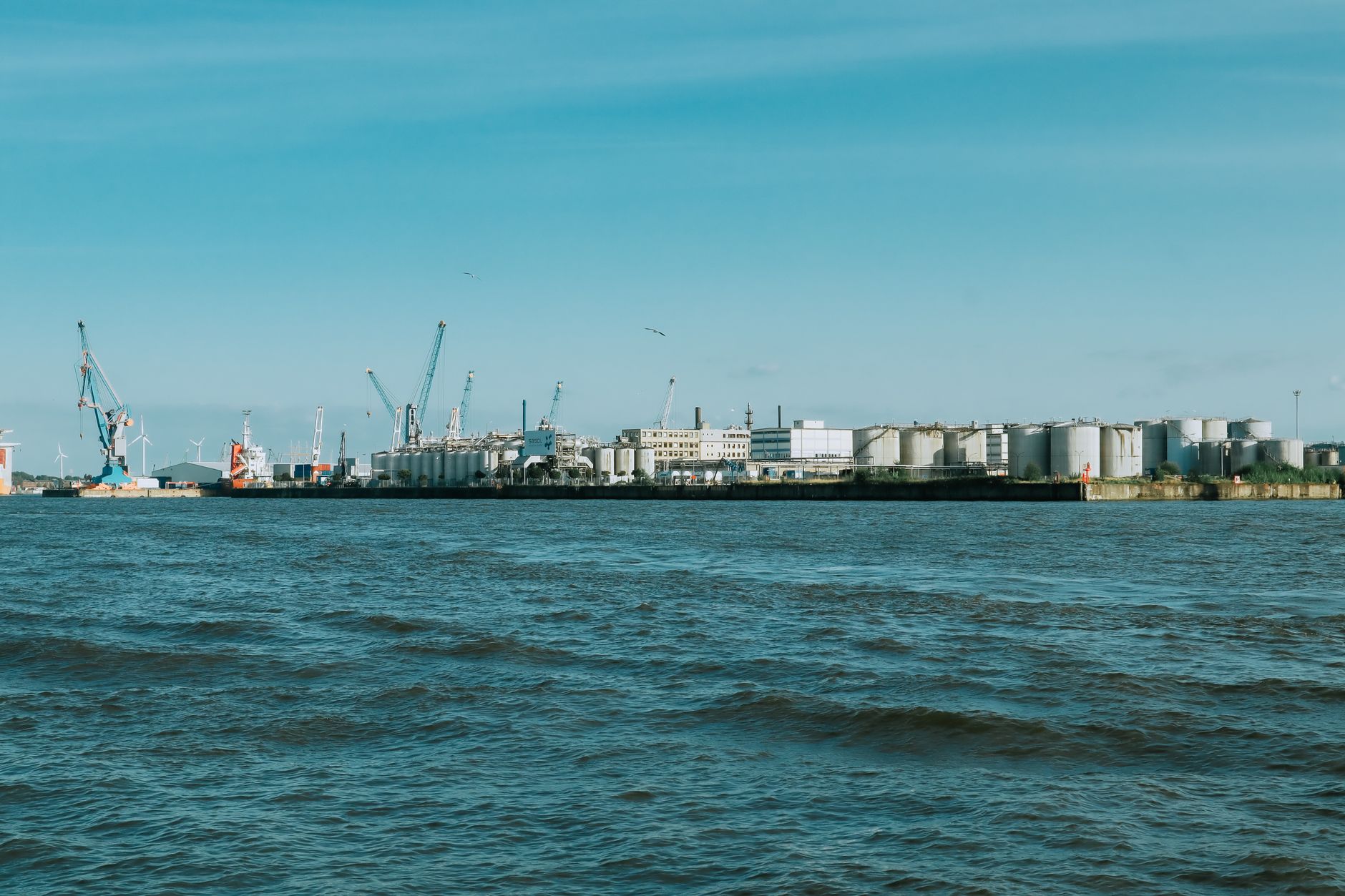 aerial view of a commercial port's oil terminal, with large cylindrical storage tanks and loading infrastructure visible along a coastline, no vessels in frame
