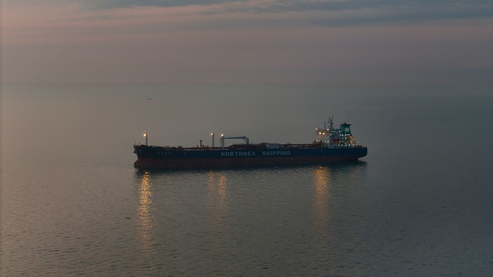 a large crude oil tanker transiting a narrow maritime strait, viewed from an elevated coastal vantage point, calm blue water with arid hillsides in the background