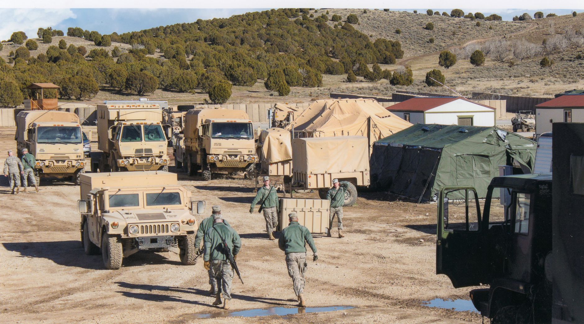rows of U.S. Army armored vehicles parked under camouflage netting at a NATO base, with maintenance crews visible in the background between vehicles