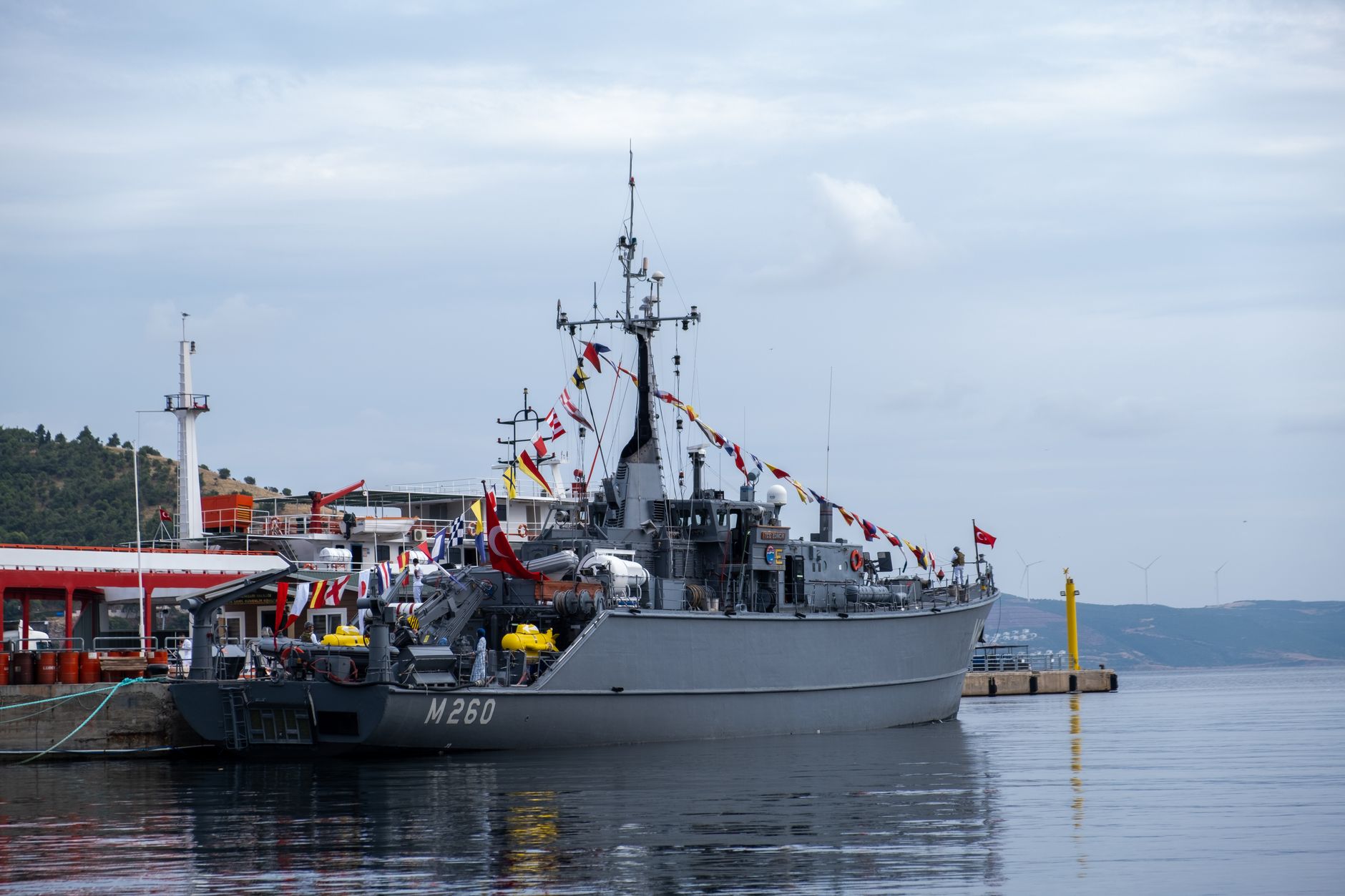 a compact remote weapon station mounted on the bow deck of a naval patrol vessel at a port facility, viewed from a low dockside angle