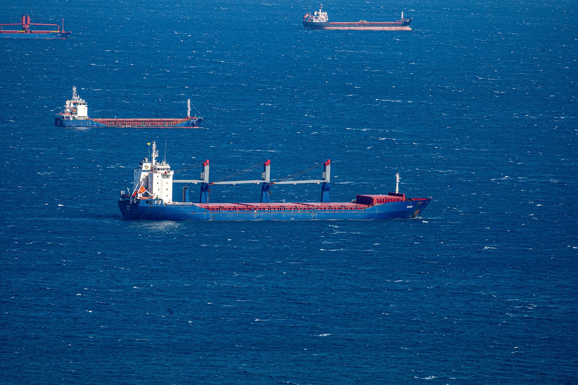 an aircraft carrier underway in open blue water, viewed from slightly above and ahead, with flight deck visible and a smaller escort vessel in the background