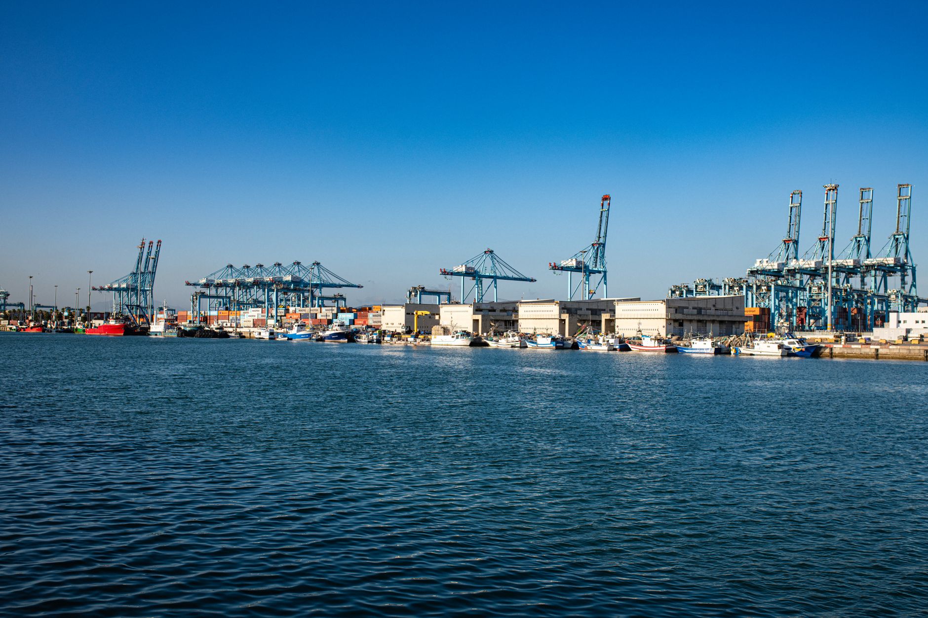 a Vanguard-class submarine moored alongside a secure Royal Navy submarine facility, with dock cranes visible in the background and calm grey water in the foreground