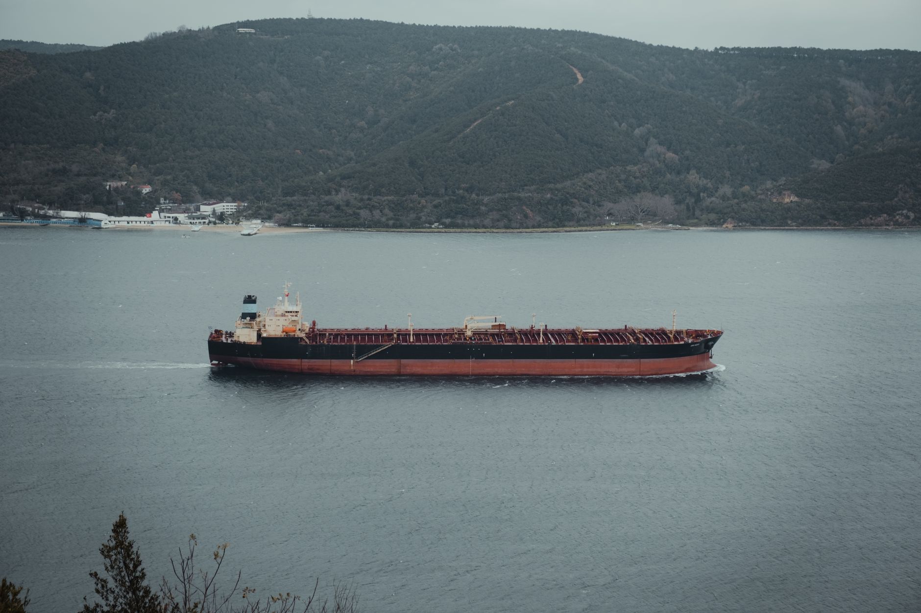 wide aerial shot of a large oil tanker navigating a coastal shipping lane near an industrial port complex, no identifying text visible
