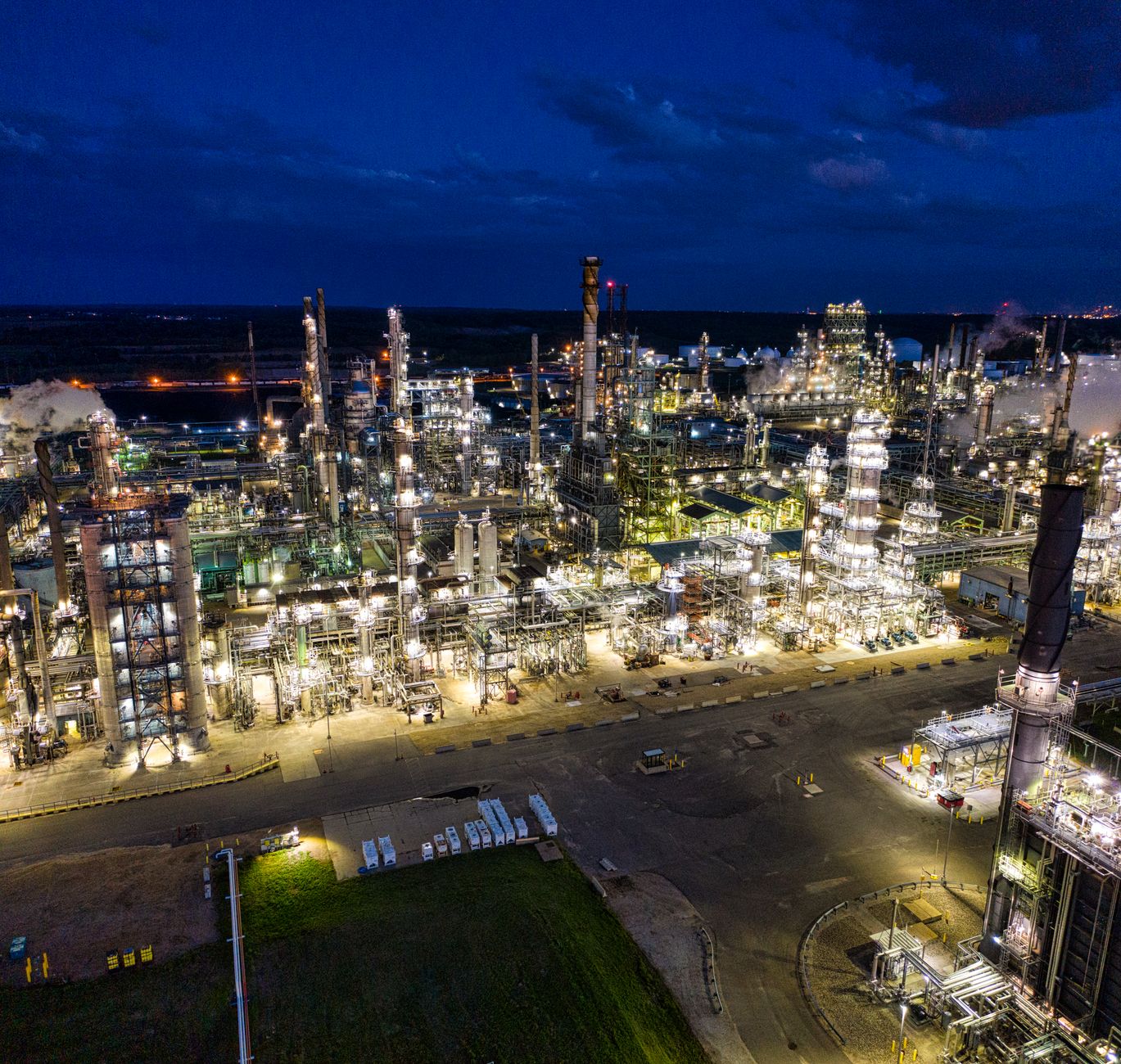 rows of industrial oil refinery distillation towers photographed at dusk, with pipeline infrastructure visible in the foreground