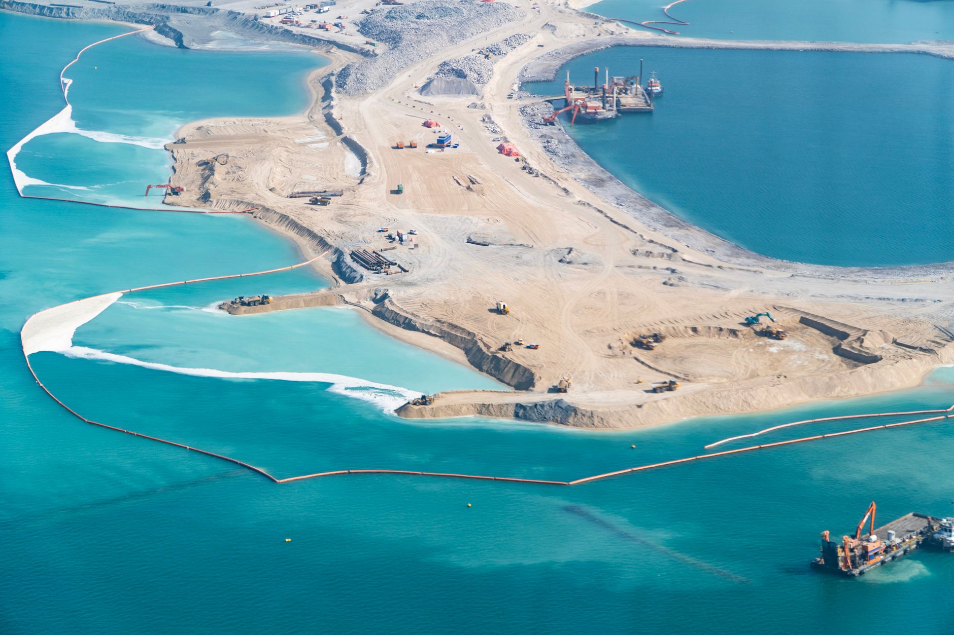 wide aerial satellite view of a newly constructed artificial island surrounded by shallow reef waters, with visible land reclamation dredging marks and early-stage infrastructure pads