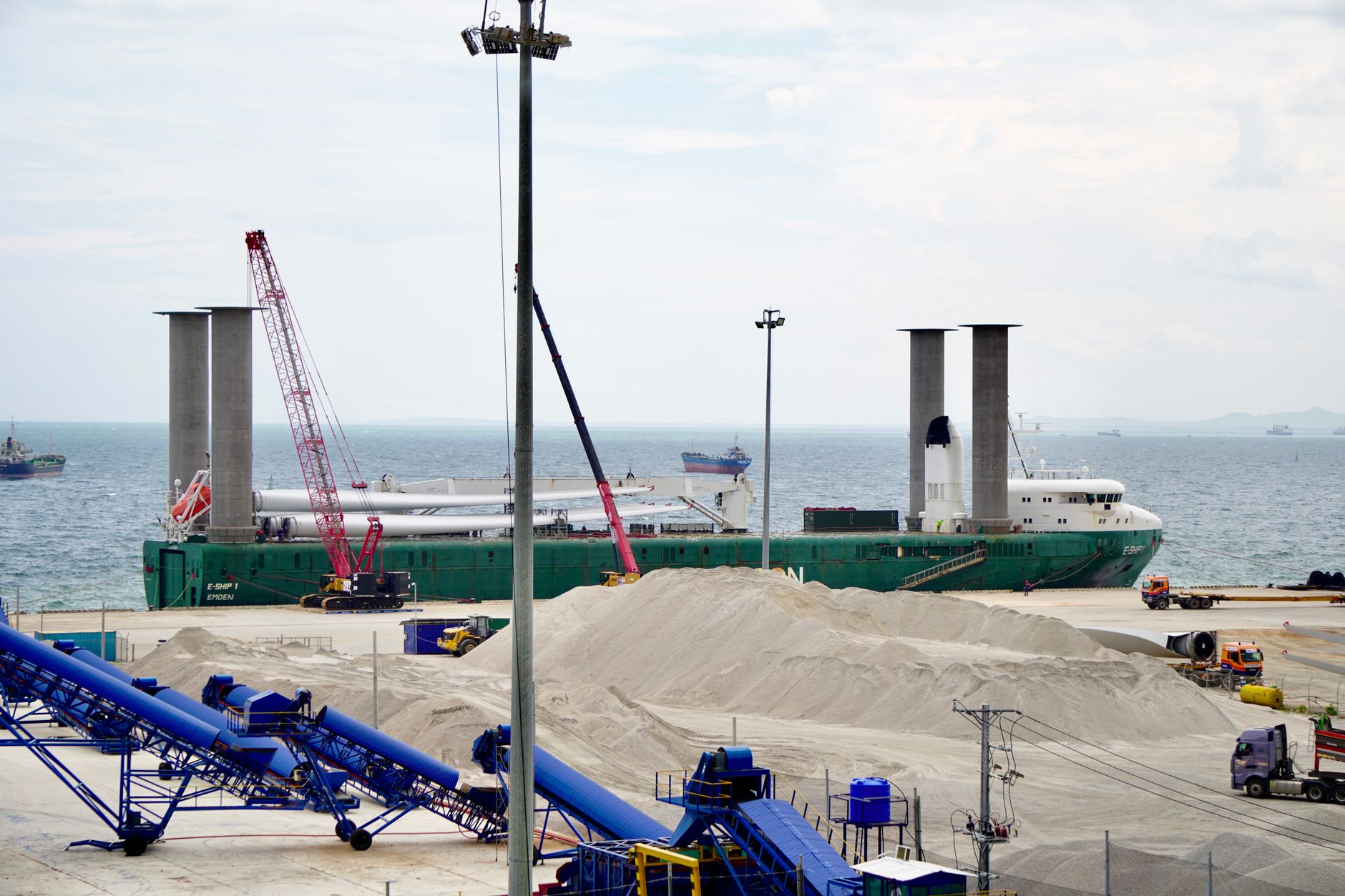 a tall reusable rocket booster standing upright on a coastal launch pad, surrounded by recovery support equipment and service vehicles, industrial port infrastructure visible in background