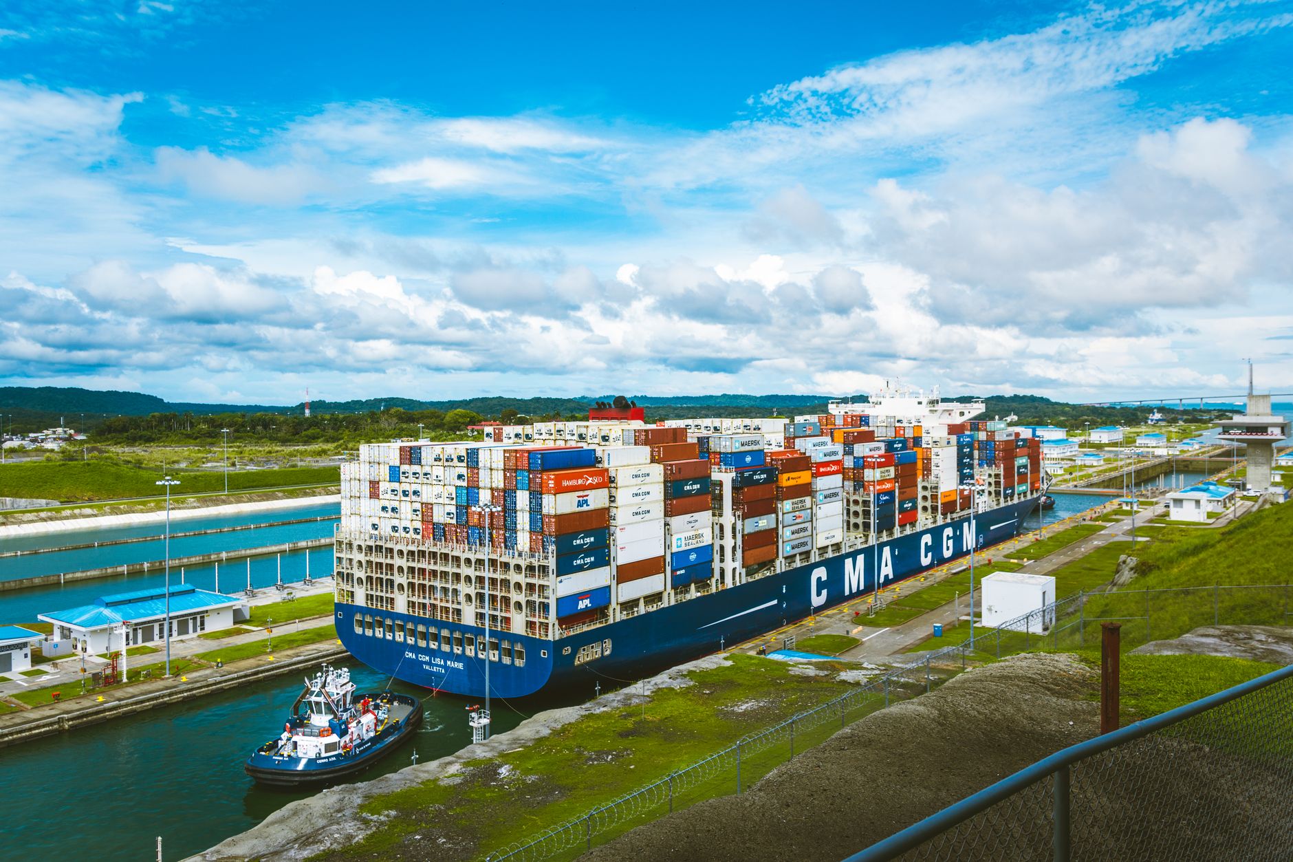 a cargo container ship transiting a narrow strait, viewed from a distance, with a naval escort vessel visible in the background