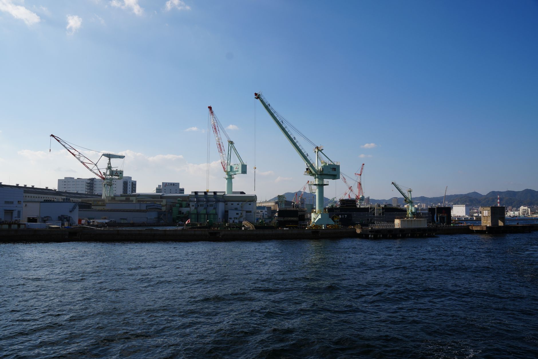 a large naval shipyard facility with dry docks and cranes visible along a waterfront, industrial buildings in the background