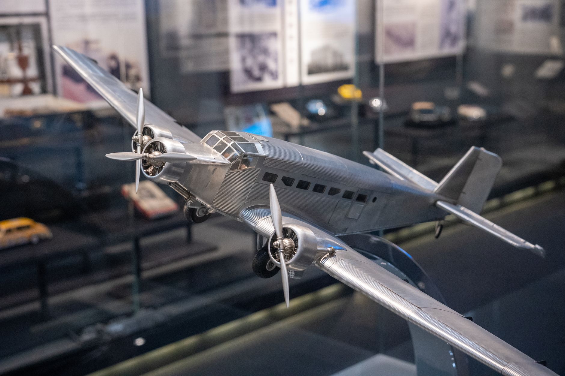 interior of a large aerospace exhibition hall with scale aircraft models and booth structures in the background, a wide exhibition floor visible with display cases and technical panels along the walls