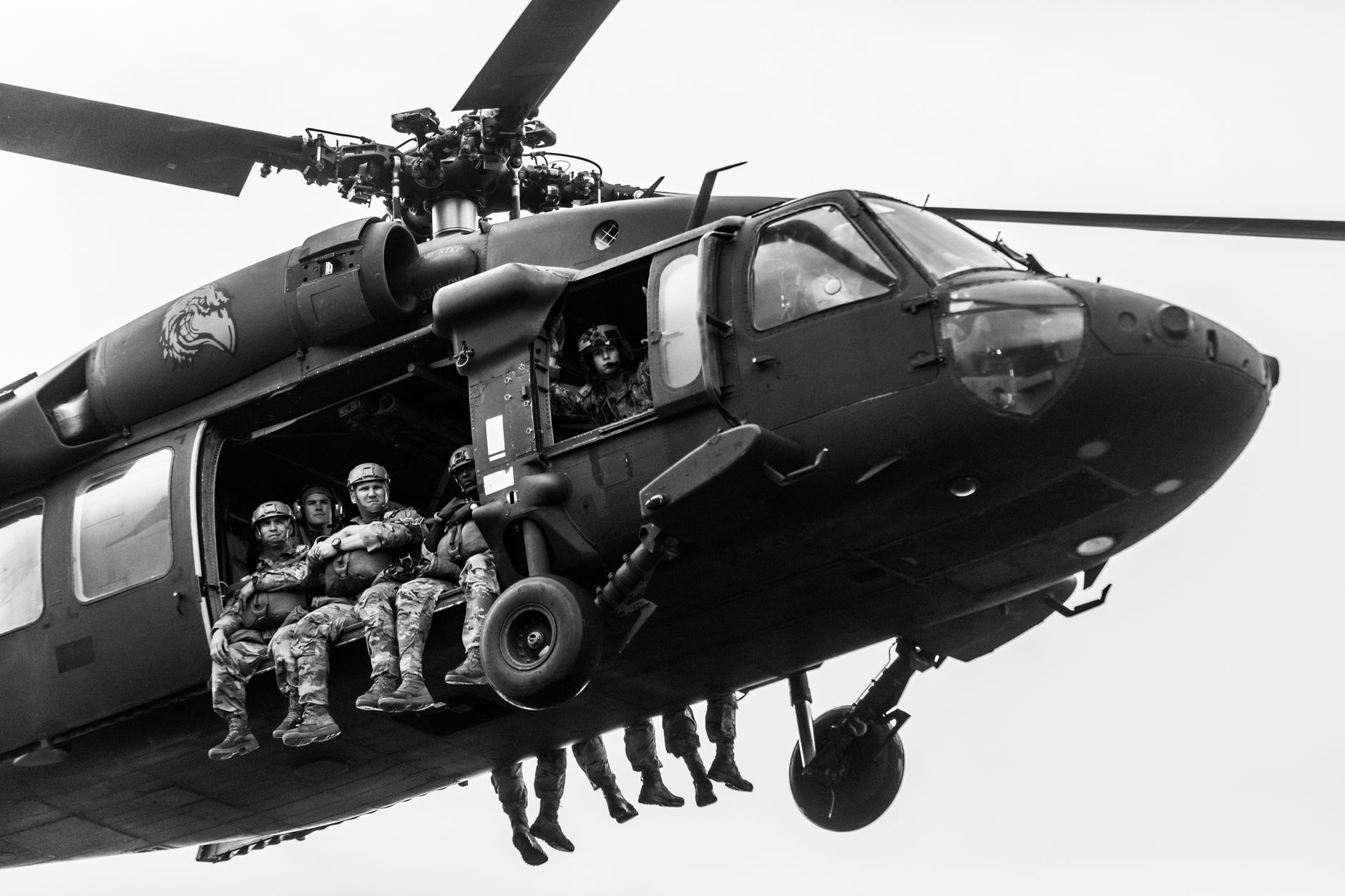 An AH-64 Apache attack helicopter parked on a tarmac at a military air base, rotor blades folded, ground crew equipment visible in the foreground