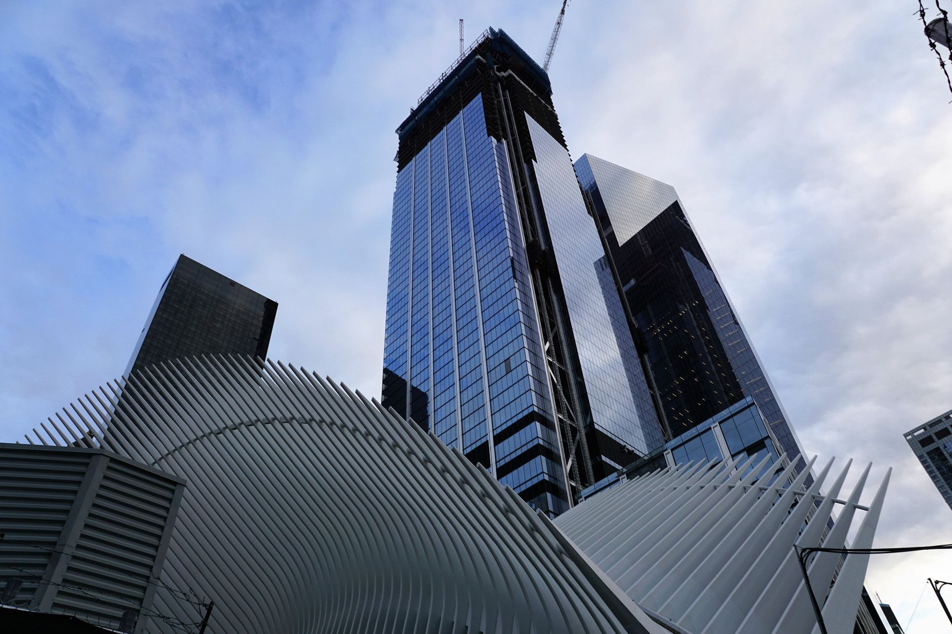 a city block in lower Manhattan with modern glass office towers, street-level storefronts, and pedestrian traffic visible from an elevated vantage point