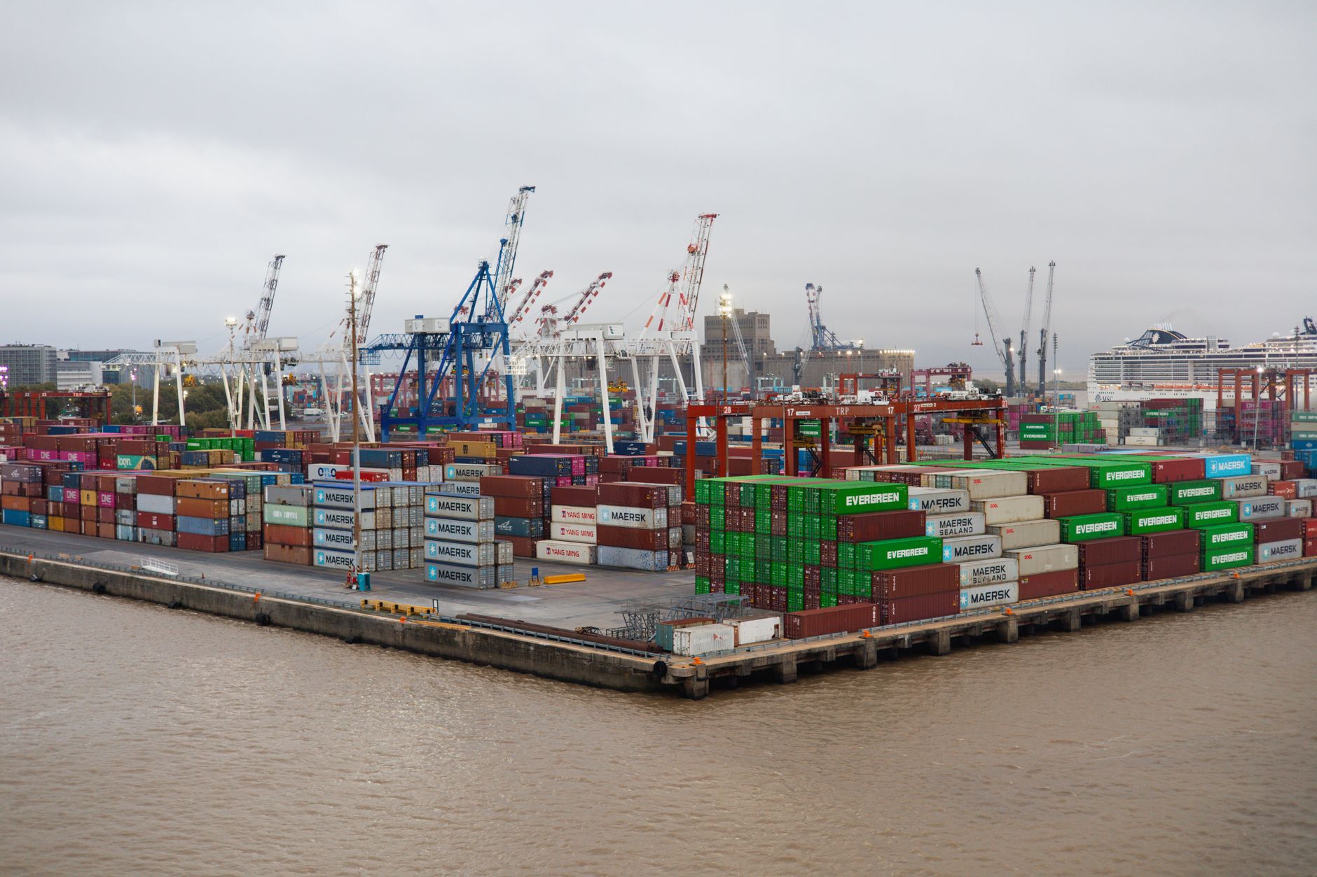a modern port facility with military cargo vessels docked alongside commercial shipping containers, cranes operating in the background under overcast skies