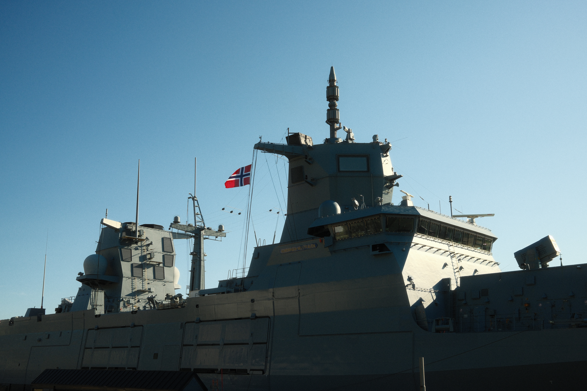 a US Navy Arleigh Burke-class destroyer at anchor in calm blue-green open ocean, viewed from a low angle slightly off the bow, no people in foreground