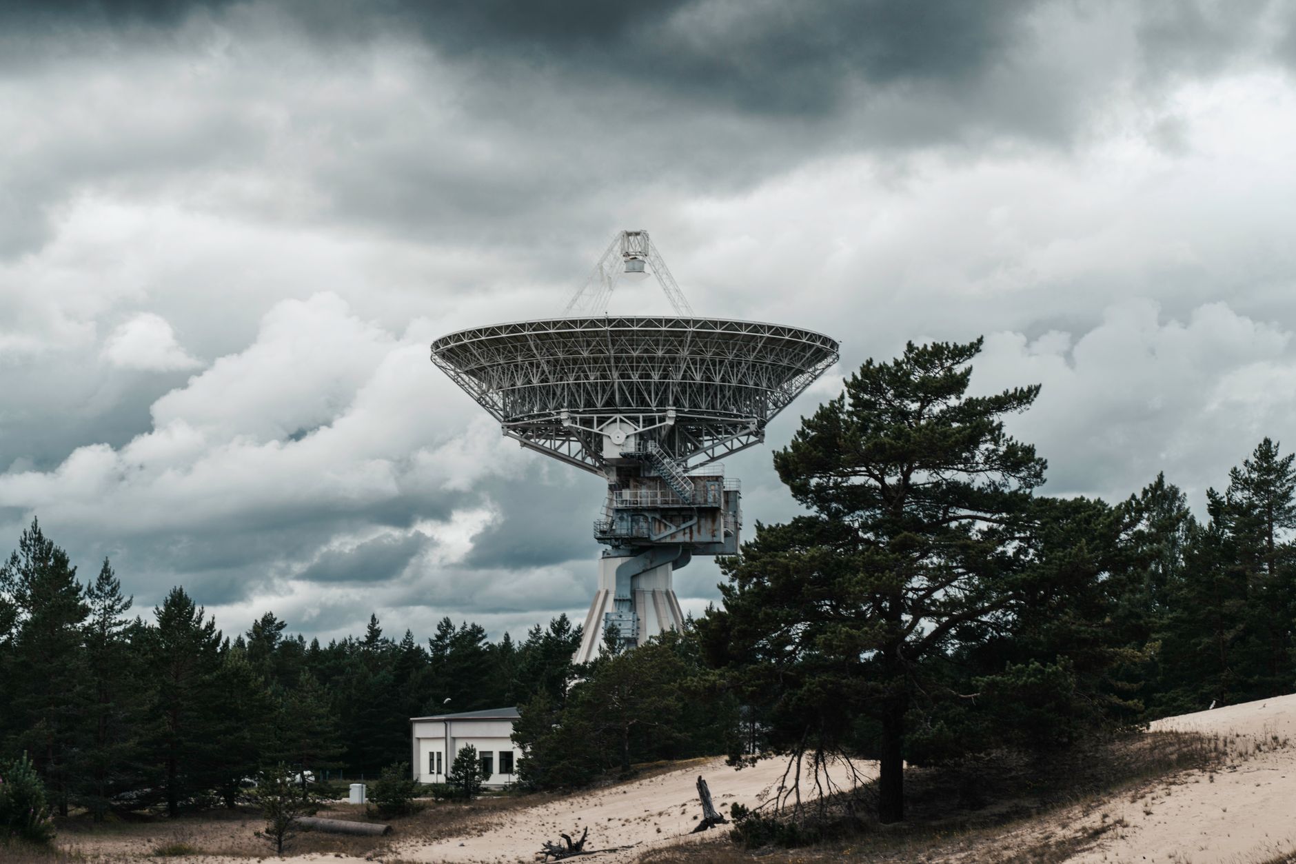 a missile defense radar installation on an open plain, its large rotating dish antenna elevated against an overcast sky with support structures and power cables visible in the foreground