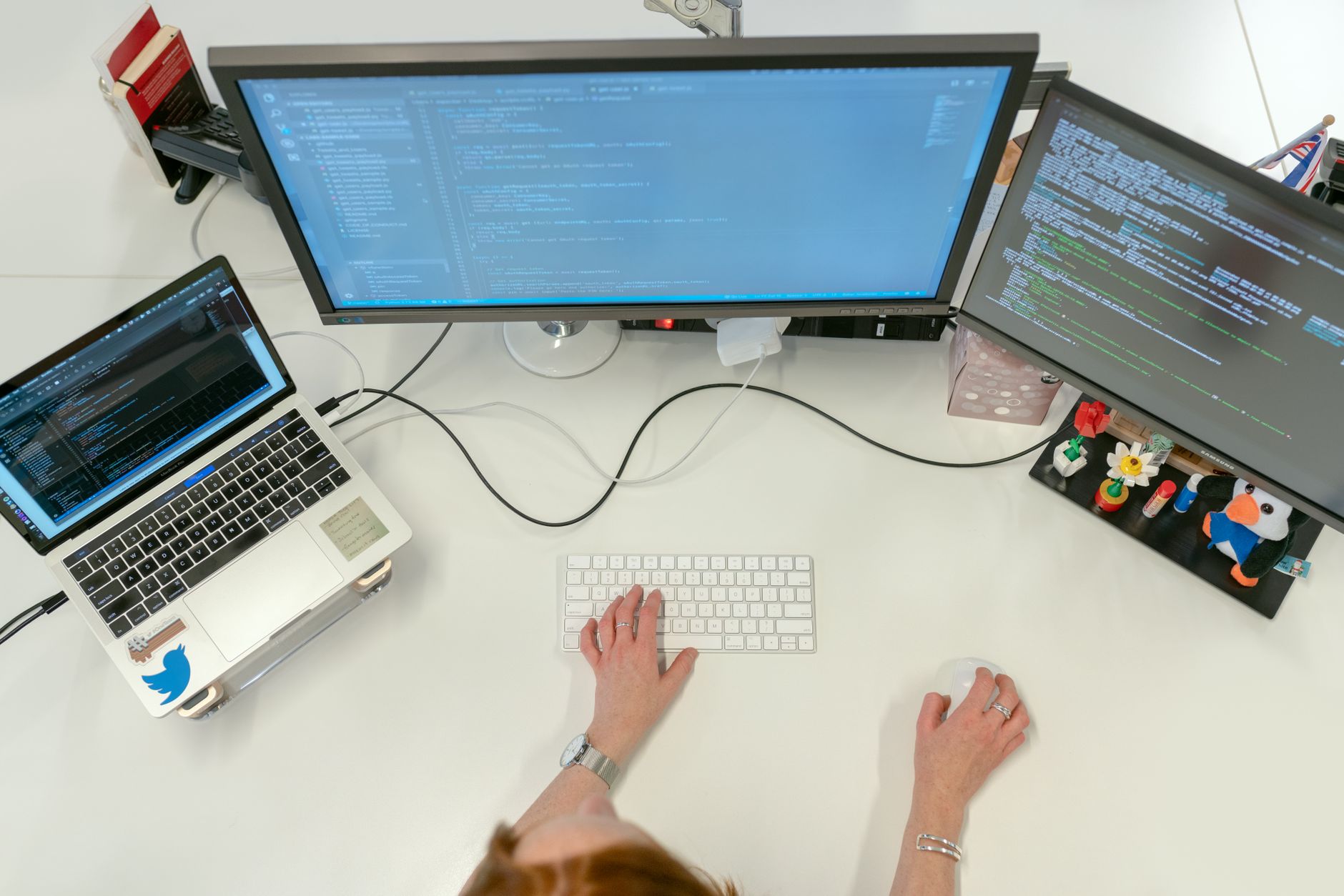 an open-plan software development office with multiple workstations displaying code editors and network monitoring dashboards, photographed from a wide overhead angle