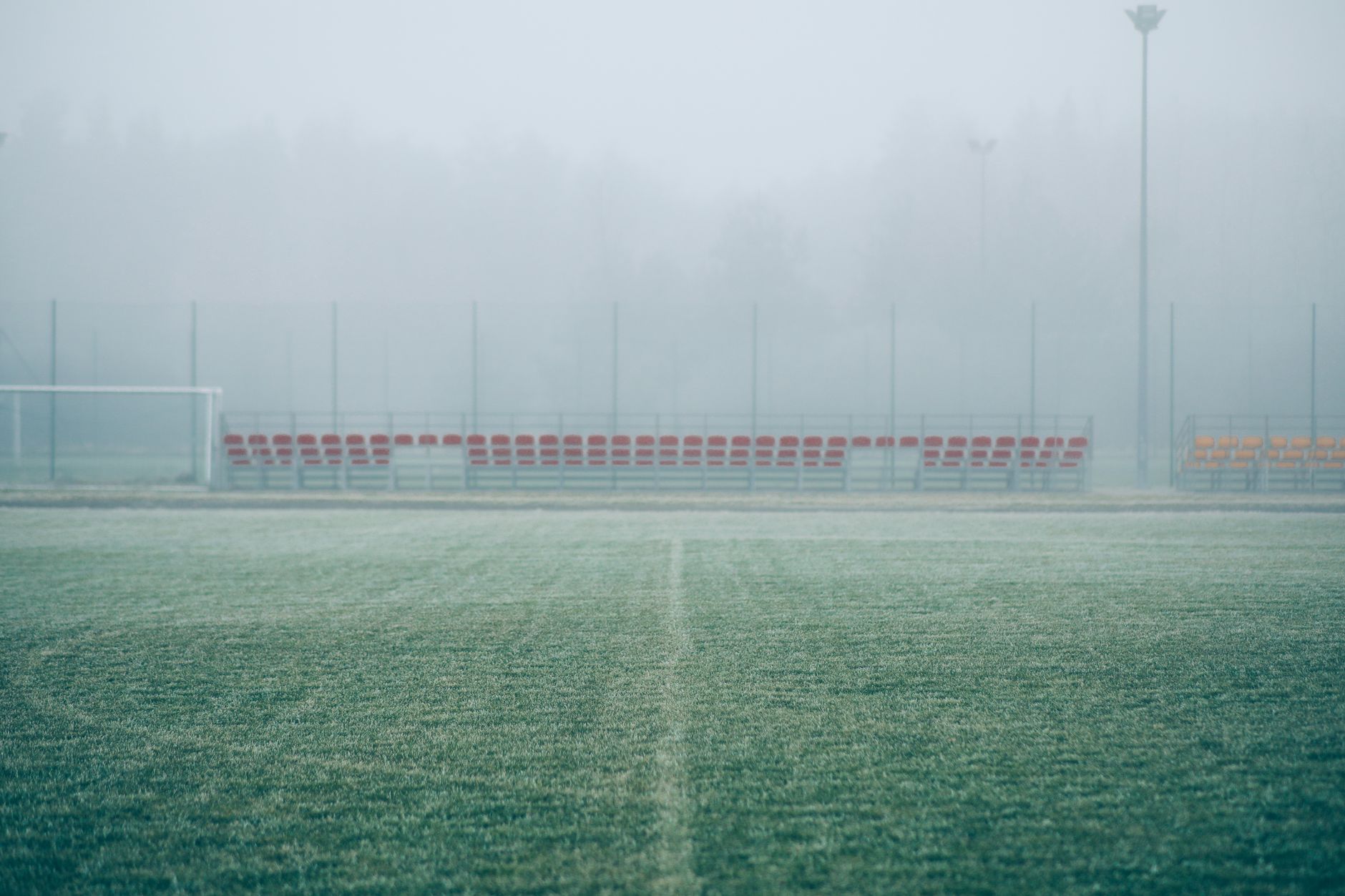 a professional football practice field with goal posts and yard line markings visible under summer sunlight, empty stadium seating in the background