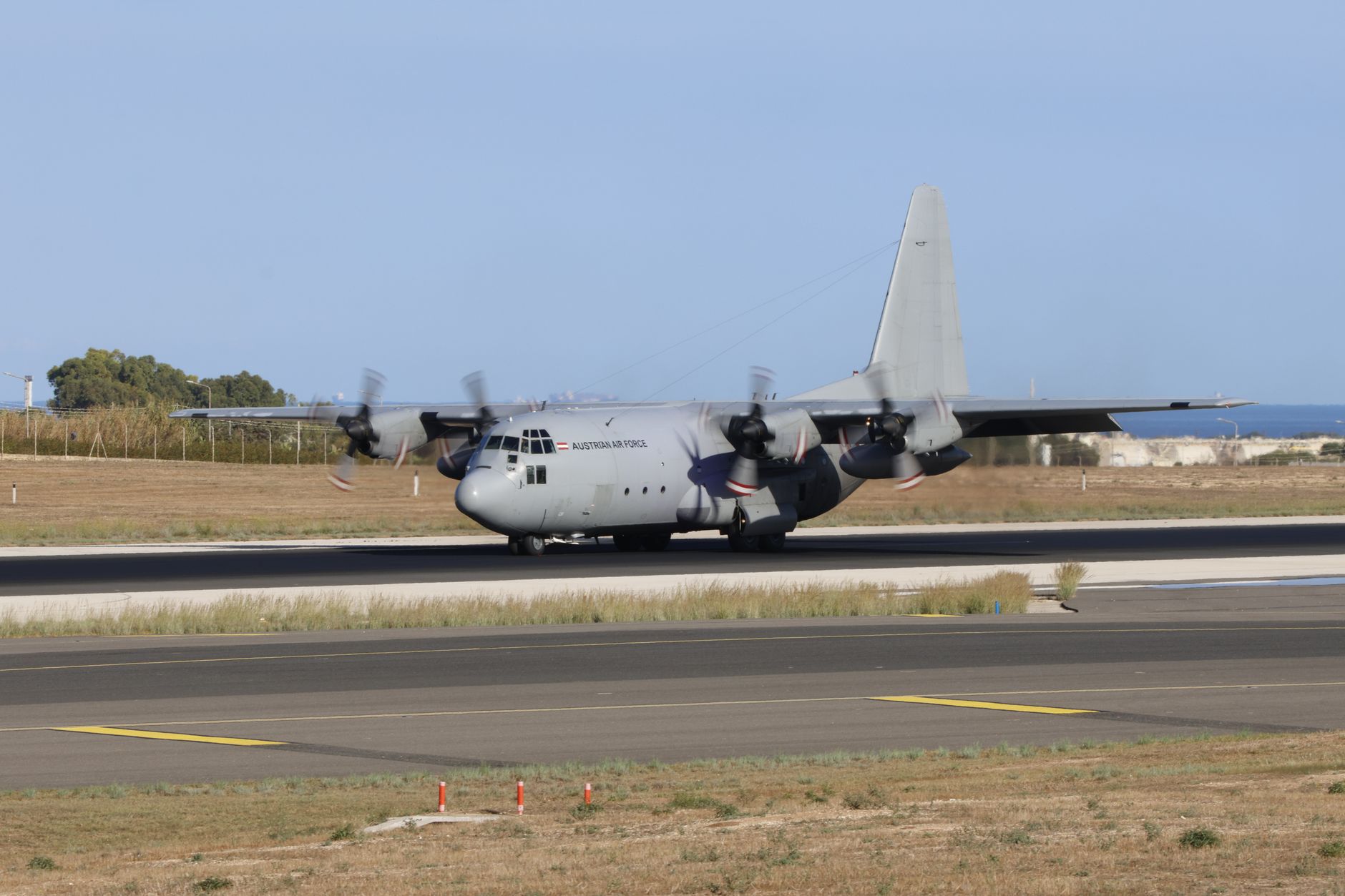a C-130 Hercules transport aircraft parked on a tropical airfield apron, ground support equipment visible nearby, dense vegetation along the runway perimeter