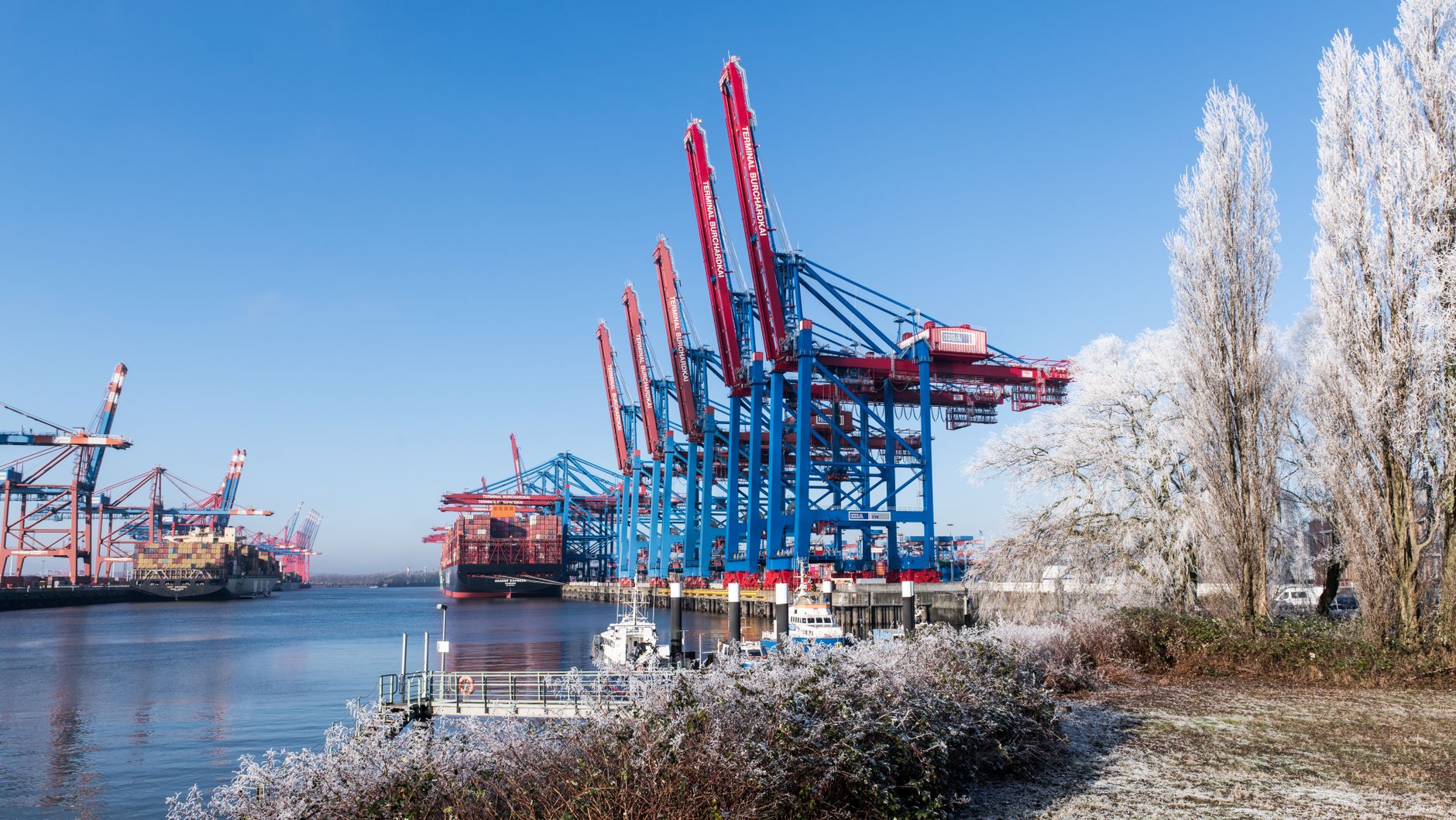 a large nuclear submarine hull being floated out from a covered dry dock at an Arctic shipyard, viewed from a wide dockside angle with cranes overhead and snow-dusted infrastructure visible in the background
