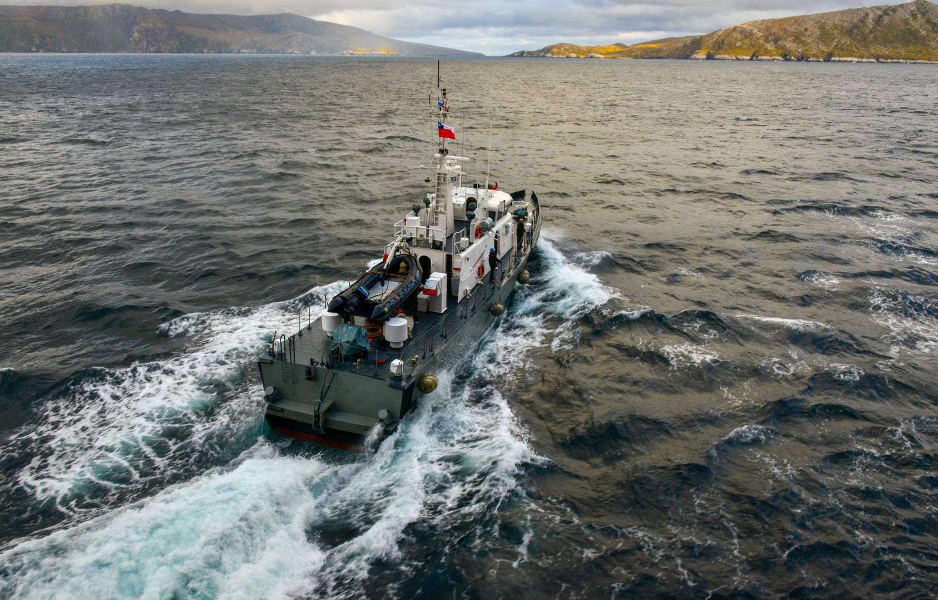 an unmanned surface vessel underway on open water near a coastal shoreline, with calm sea conditions and distant hills visible in the background