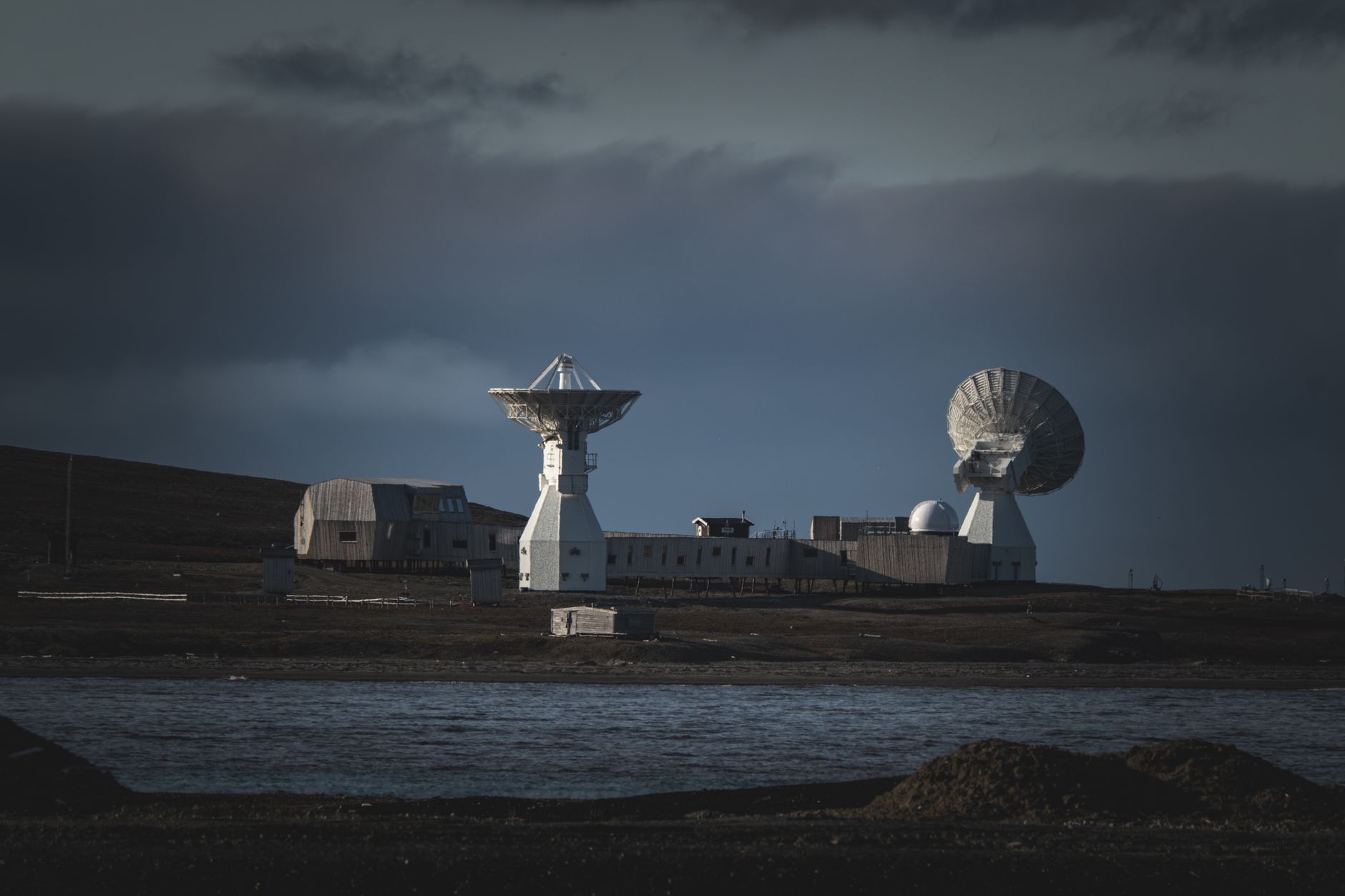 an outdoor satellite ground station at dusk, showing large parabolic dish antennas pointed skyward against an overcast sky, surrounded by security fencing and low support buildings