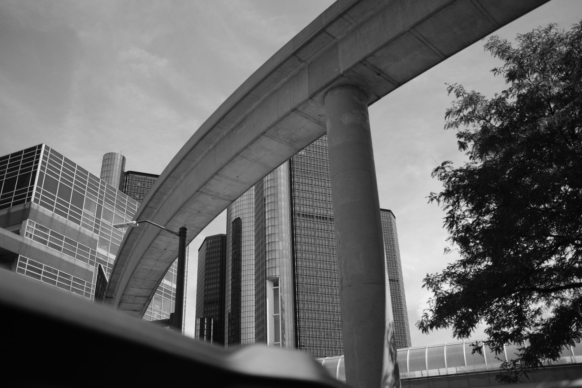 An M1A2 Abrams main battle tank parked beneath a low concrete overpass in a dense urban district, surrounded by civilian commercial buildings, viewed from a wide street-level angle