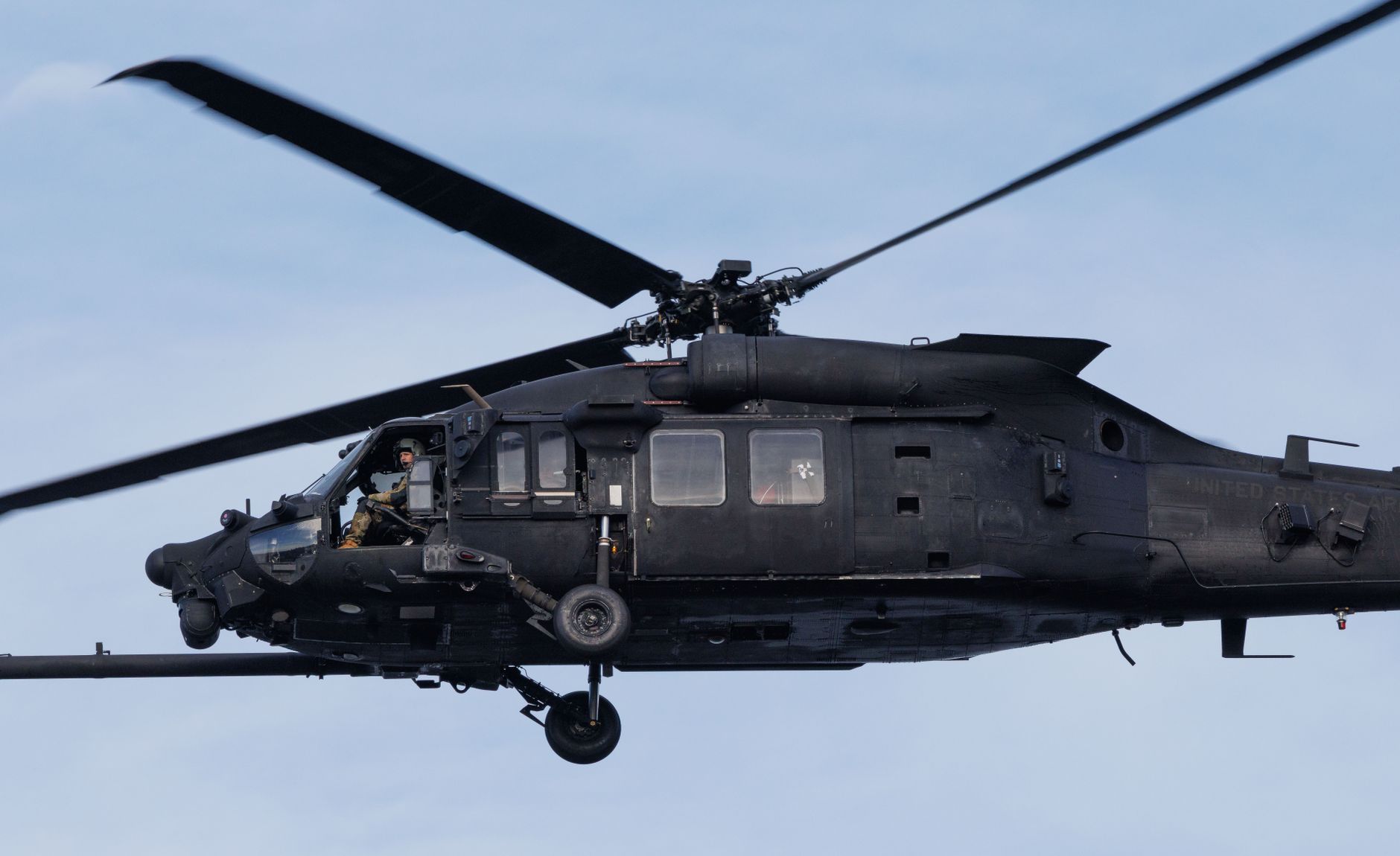 an AH-64 Apache attack helicopter on a flight line at a military air base, viewed from the side with mountains visible in the background