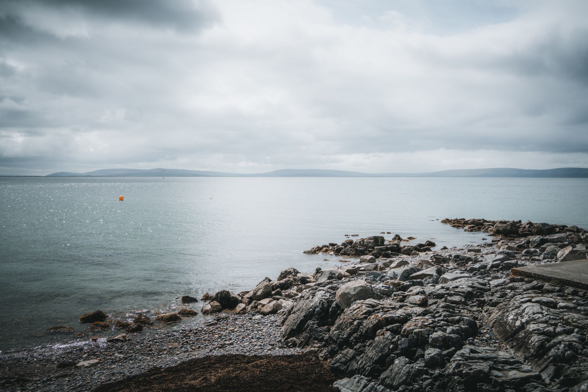 a mobile military launcher vehicle positioned along a rocky coastline, with the sea visible in the distance and overcast sky above