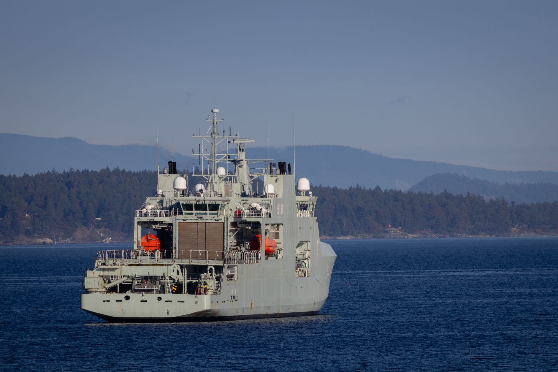 a naval frigate underway during sea trials, viewed from a low angle at the stern with a calm open sea in the background and visible launch system cells on the foredeck