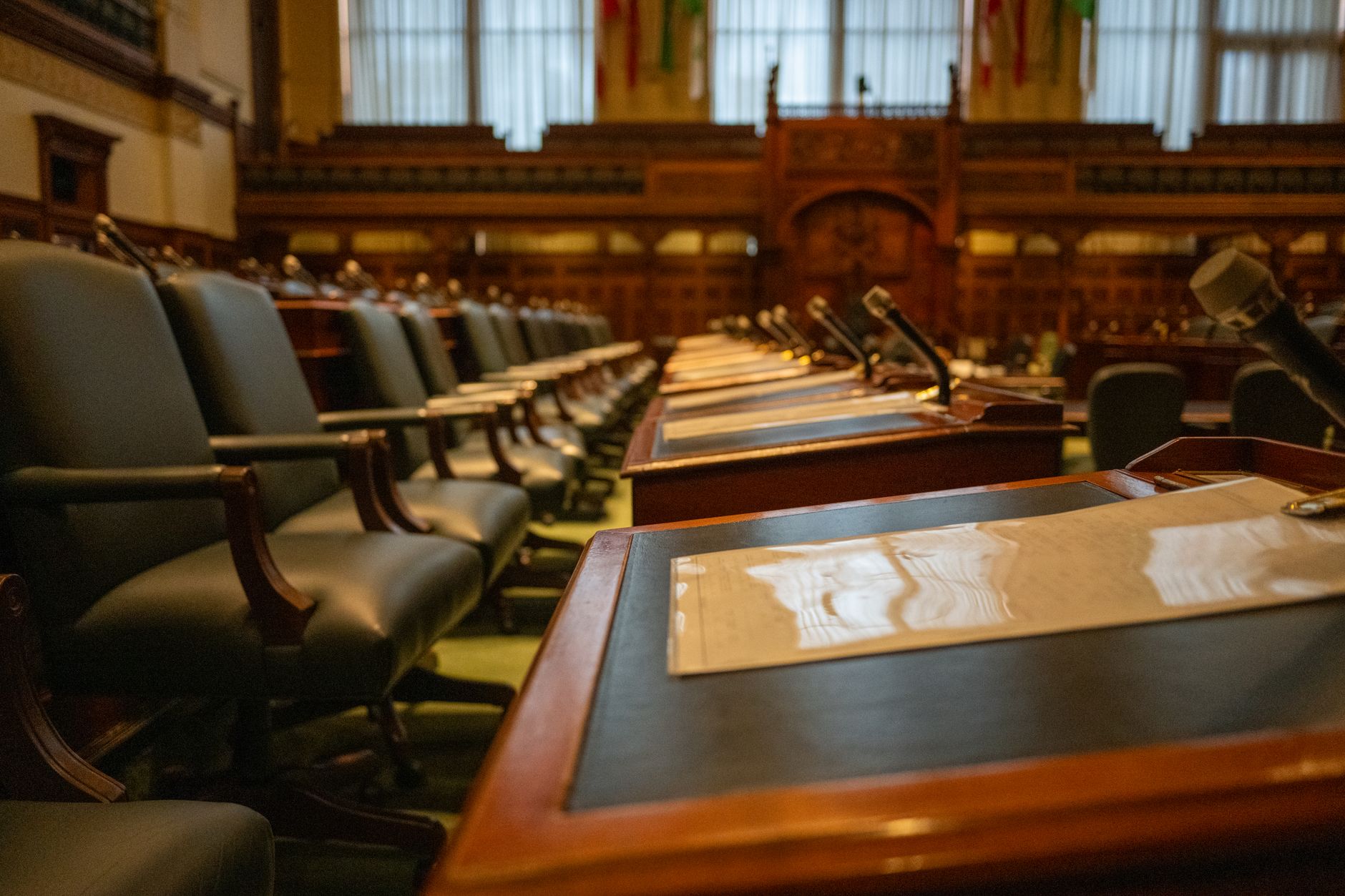 a long wood-paneled federal hearing room interior, empty leather chairs at a curved dais with microphones, American flags on either side of a central seal on the wall