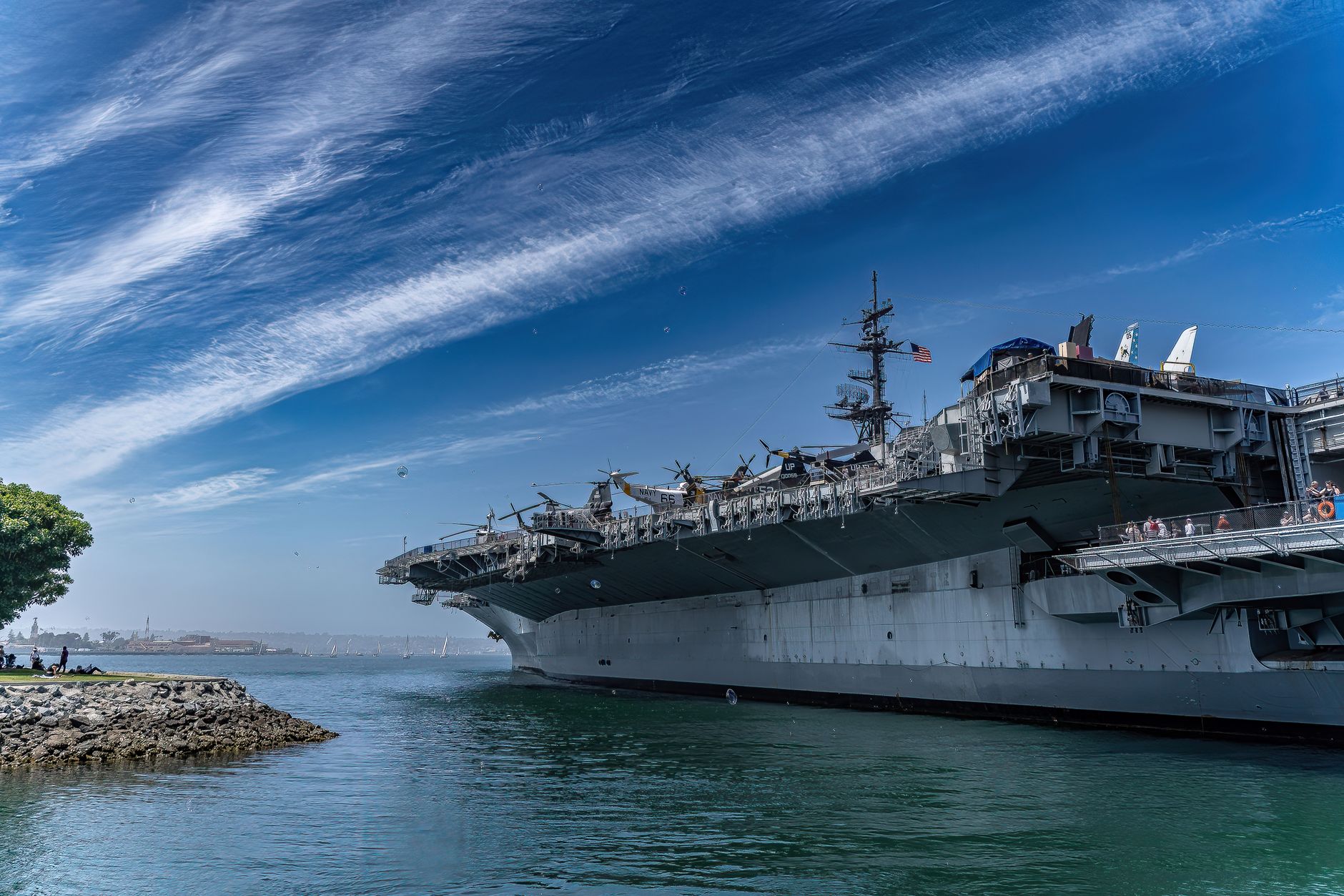 a Nimitz-class aircraft carrier in dry dock at a naval shipyard, with scaffolding along the hull and maintenance equipment visible on the surrounding pier