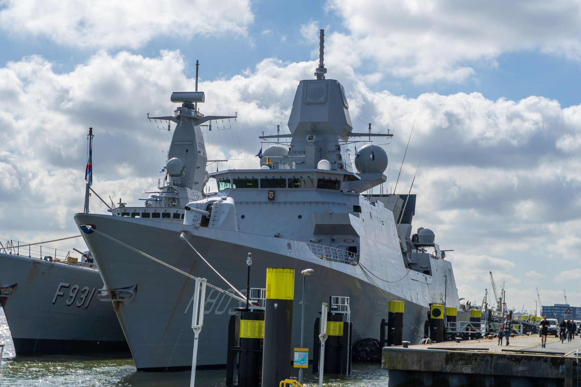 an Arleigh Burke-class guided-missile destroyer moored at a naval station pier, viewed from the dock with its hull and superstructure in full profile against an overcast sky