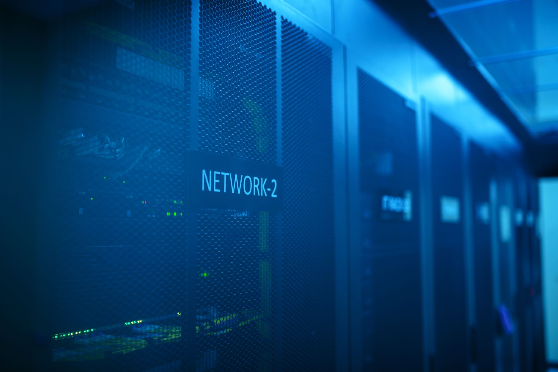a server room with rows of illuminated rack-mounted hardware inside a modern data center facility, shot from a wide angle showing the length of the aisle