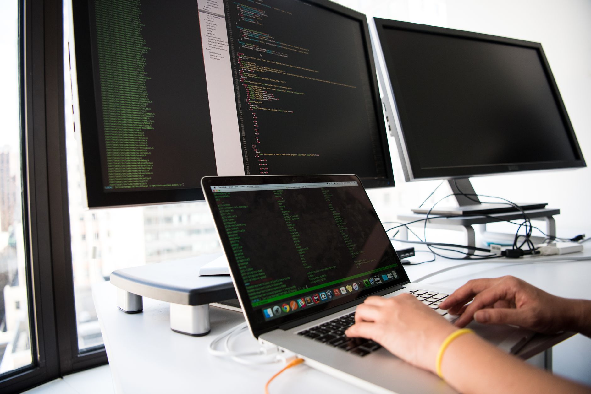 a software engineer's workstation displaying multiple open monitoring dashboards with network flow diagrams and alert logs on dual screens, in a dimly lit office environment