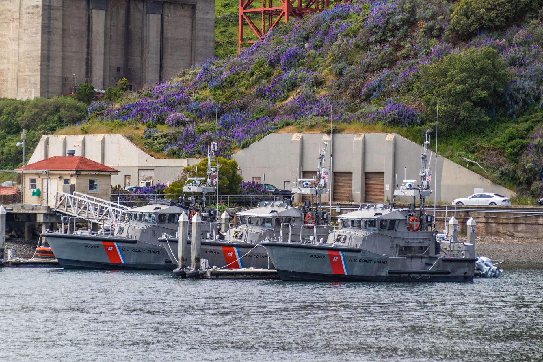 a naval dockyard facility with patrol vessels moored alongside maintenance infrastructure, cranes visible in the background
