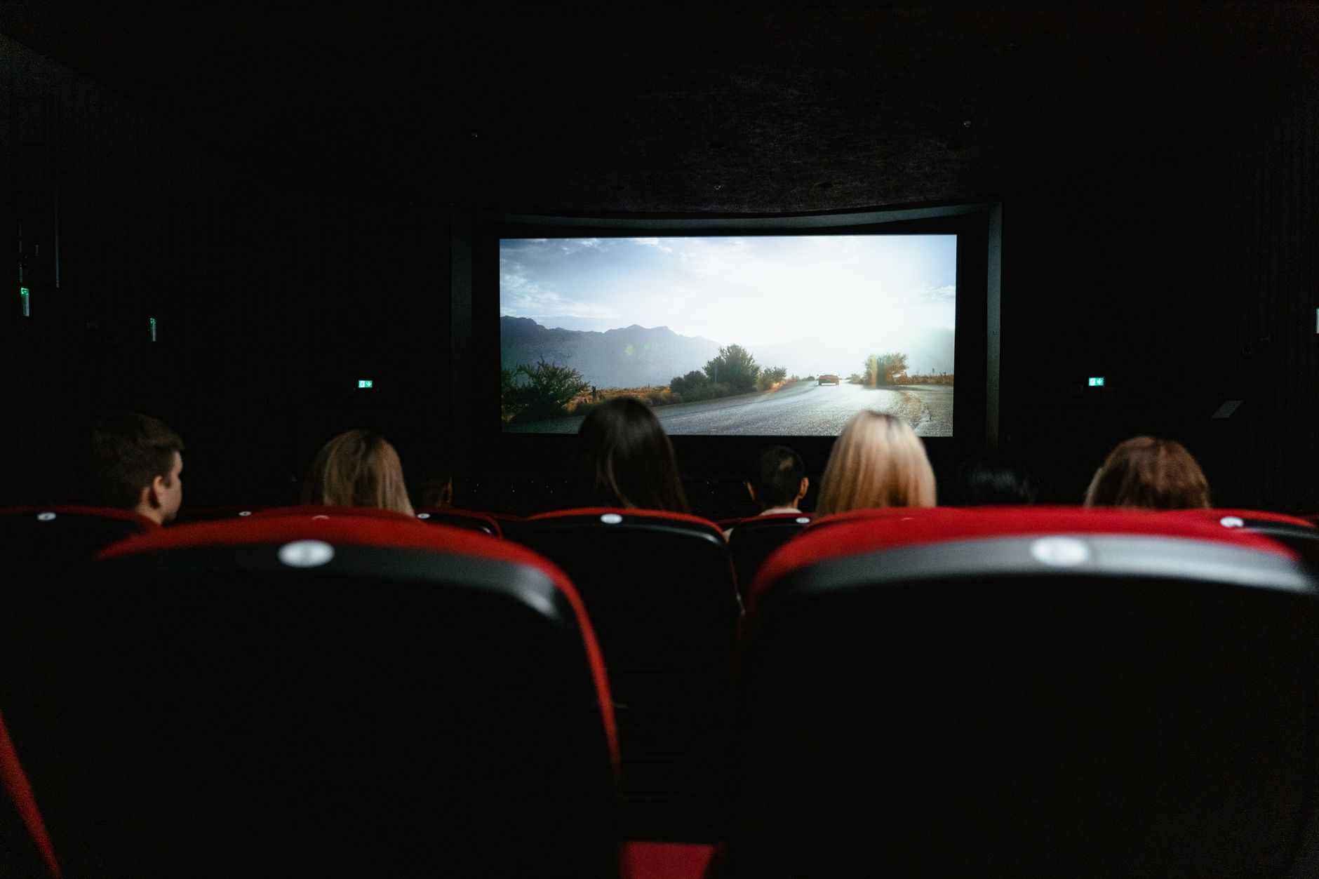 an empty screening room with rows of seats facing a large projection screen showing a black-and-white WWII-era landscape still frame, ambient light from ceiling fixtures