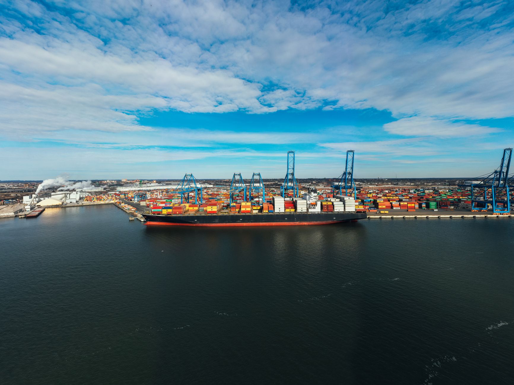 a wide aerial view of a busy East Asian naval base with multiple vessels at dock, cranes active over a large gray hull under final outfitting