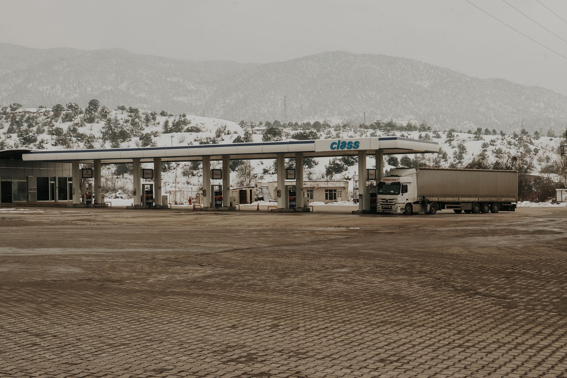 a row of long-haul diesel freight trucks parked at a highway fuel station, pumps visible in the foreground, overcast sky above an open interstate landscape