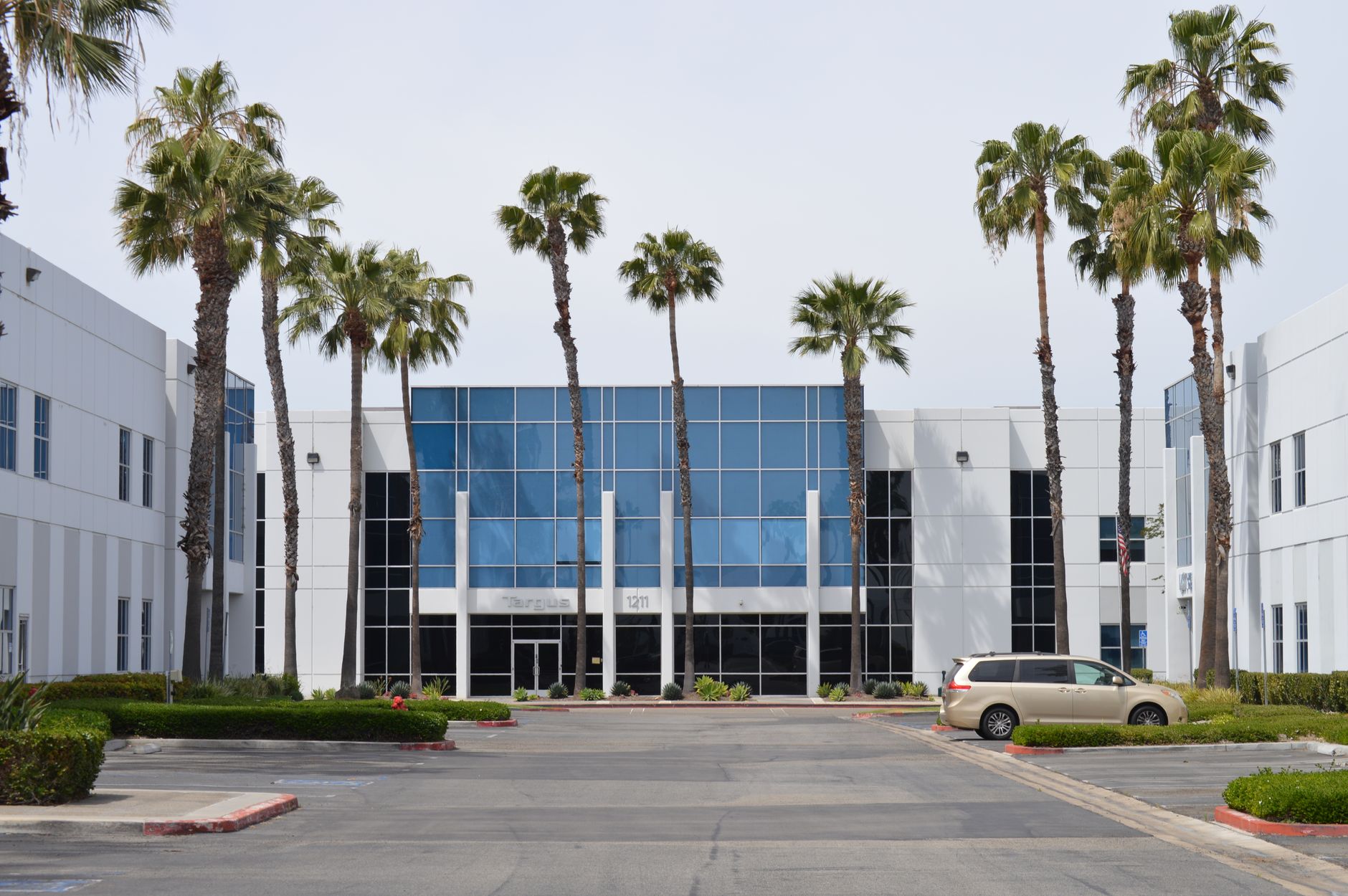 exterior of a mid-size defense contractor campus in a Texas business park, glass-facade office building with a manicured entrance, American flag mounted at the entrance, midday lighting