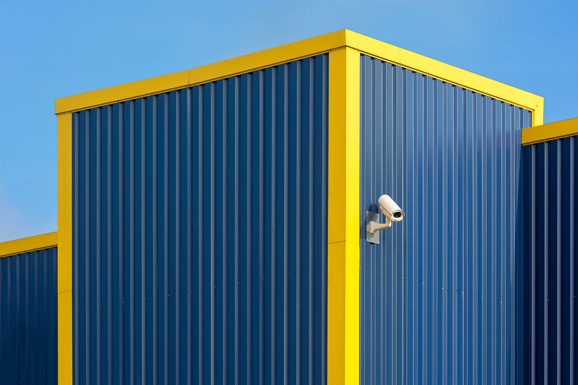 exterior of a large European defense manufacturing facility with security fencing and surveillance cameras visible along the perimeter, overcast sky