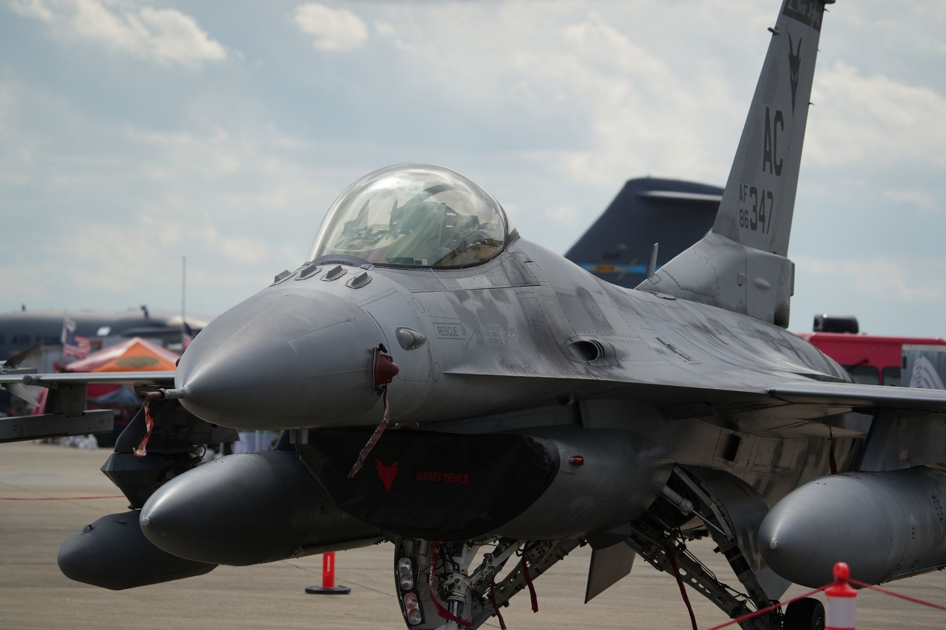 rows of F-16 aircraft parked on an outdoor flight line at a military air base, wide shot showing the tarmac and surrounding facilities