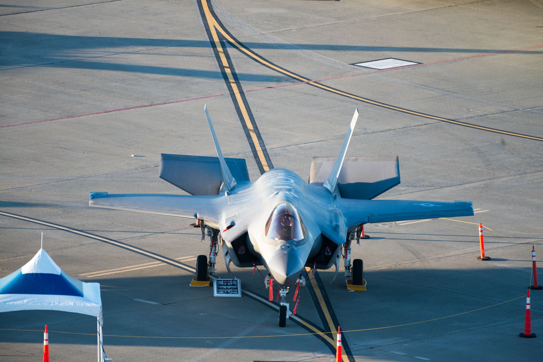 rows of F-35 aircraft components arranged on the production floor of an aircraft assembly facility, with industrial cranes and tooling visible overhead