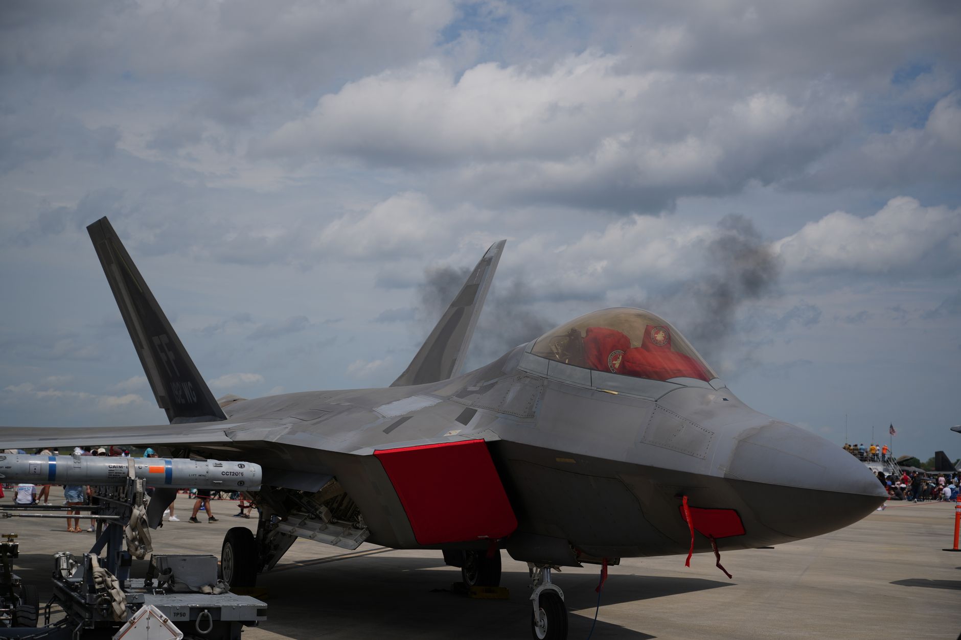 an F-22 Raptor on a flight line at a U.S. air base, viewed from a low angle with engine exhaust visible behind the aircraft against an overcast sky