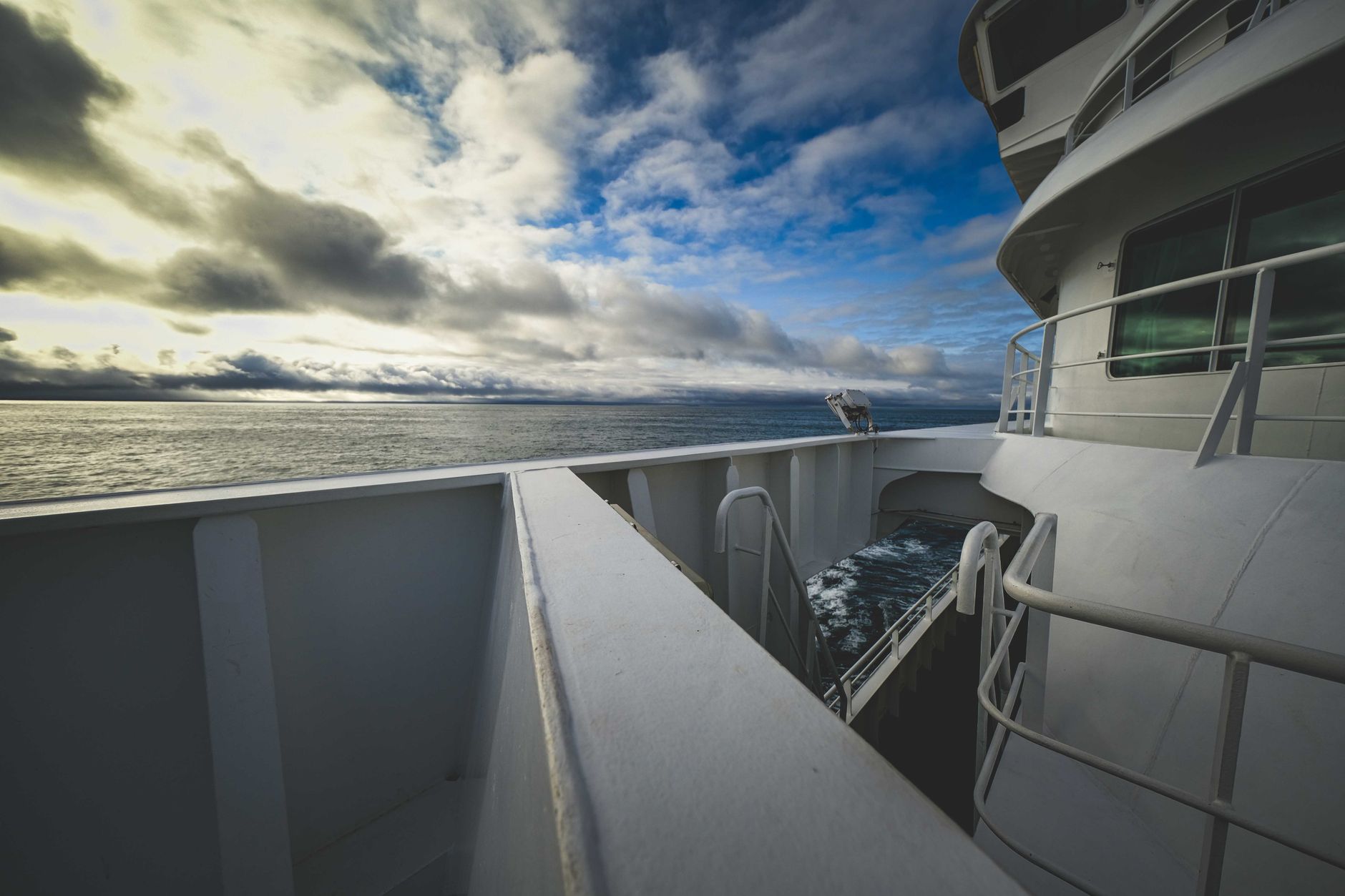 a medium-sized unmanned surface vessel underway on open ocean, viewed from a low angle from the side, with a grey hull and sensor mast visible against a partly cloudy sky