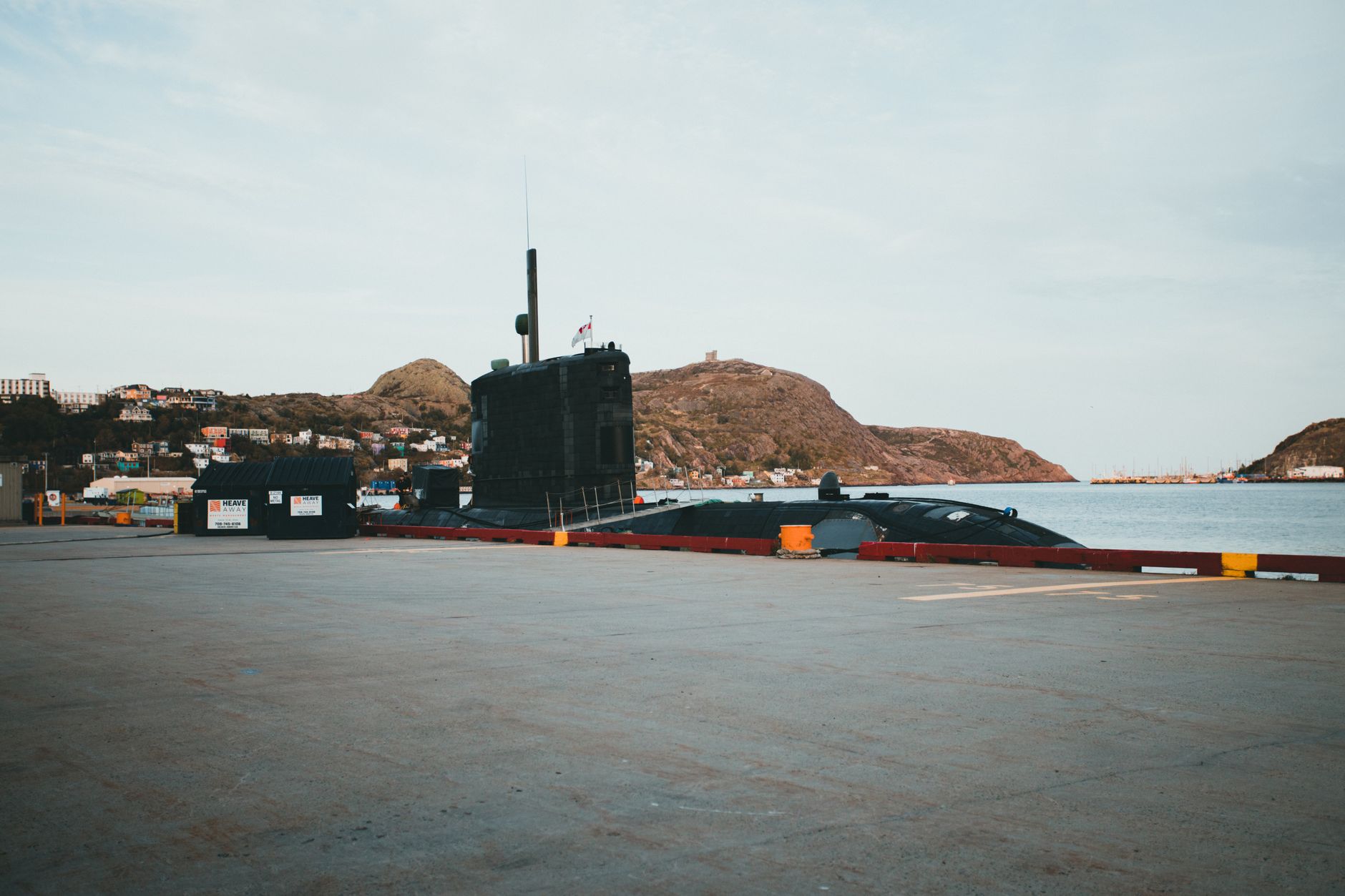 a submarine conning tower and forward hull section viewed from dockside, with open ocean visible in the background through a harbor entrance