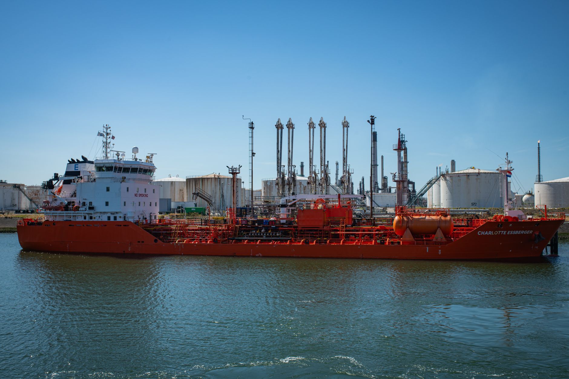 rows of oil storage tanks at a large industrial port facility seen from an elevated distance, with tankers visible at the dock