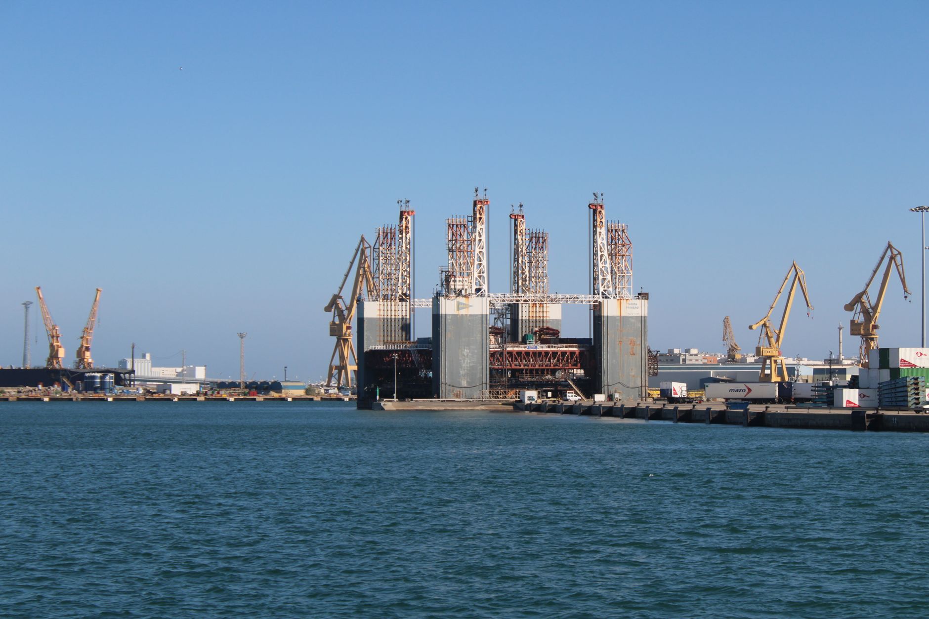 a modern diesel-electric submarine moored at a large European naval shipyard, seen from the pier with cranes and industrial infrastructure in the background