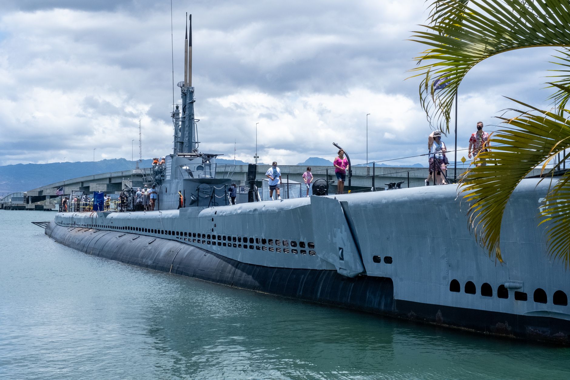 a submarine partially surfaced in calm harbor waters near a naval base pier, with support vessels visible in the background